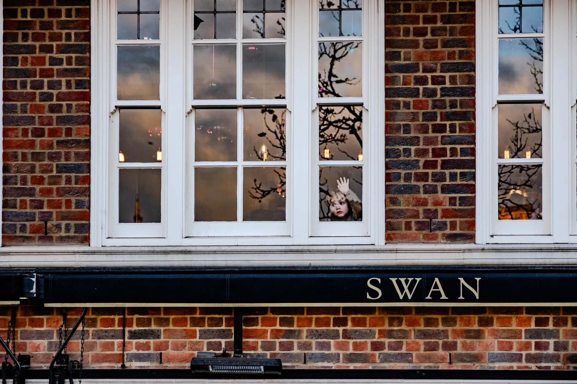 The image shows a white-framed window set into a red brick wall. The window has multiple panes and is divided into two sections. The section on the left is reflecting a cloudy sky with a white building in the distance. The section on the right shows the silhouette of a tree against a cloudy sky. A young child with blonde hair is looking out of the window, holding one arm up in the air. The wall below the window has a black awning with the word "SWAN" written in gold letters. Below the awning, the brick wall continues. There is a small metal light fixture in the lower left corner of the image.  The image appears to be of a building on a street.