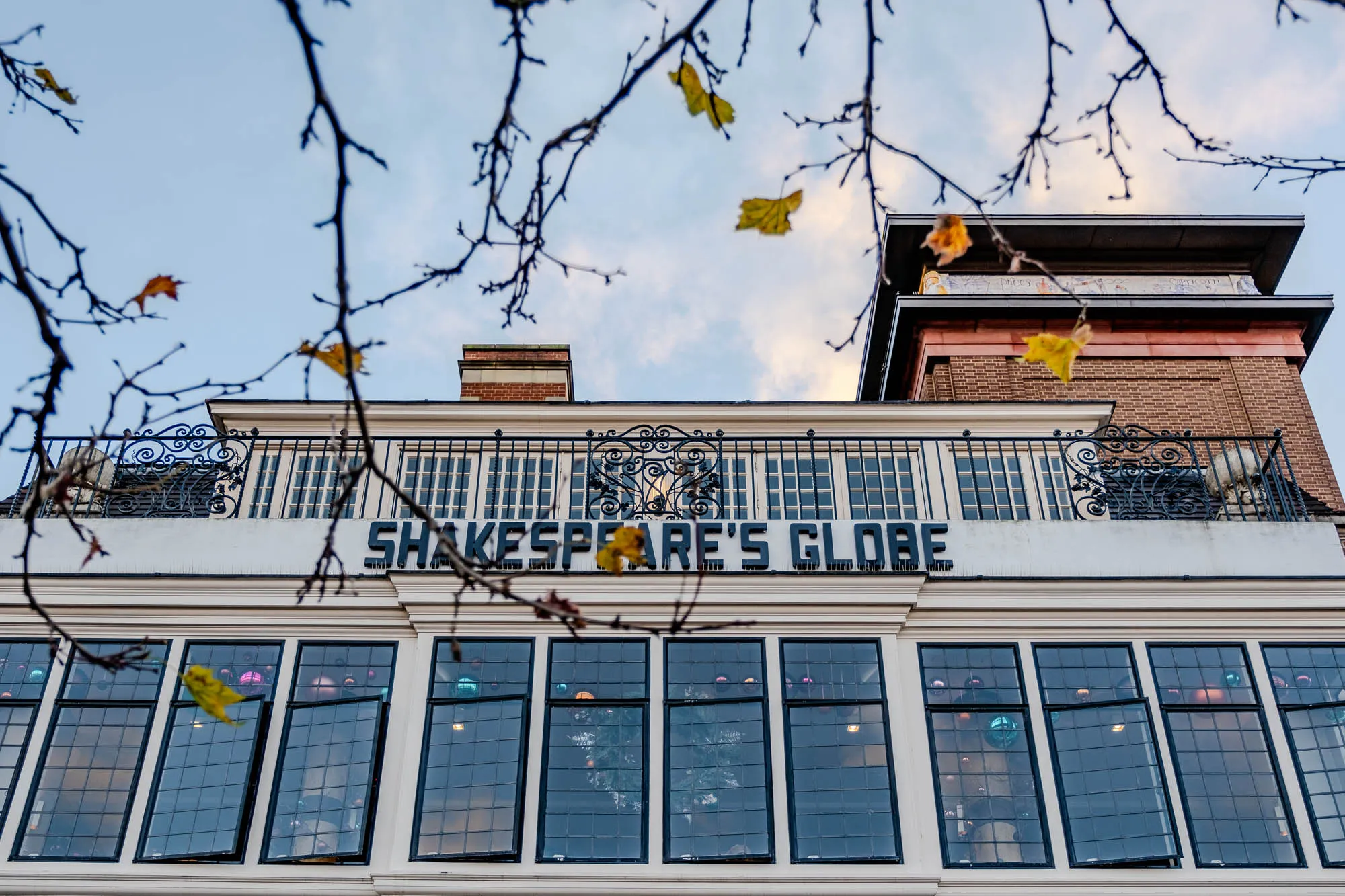 The image shows a building with the words "Shakespeare's Globe" in large lettering across the front. The building has a white facade with black trim and a black wrought iron balcony.  The balcony has a few small windows with black muntins. There are four large, tall windows with black muntins and a window sash that is open to the top.  The windows have a view of a room inside with some christmas ornaments hanging.   There is a branch with yellowed leaves reaching down from the top of the image.  Above the windows and balcony, there is a flat roof with a brown brick facade.  The sky above is blue and light pink with some white clouds.  There is a sign in the top left corner of the image.  The scene suggests an older building that is an iconic landmark.