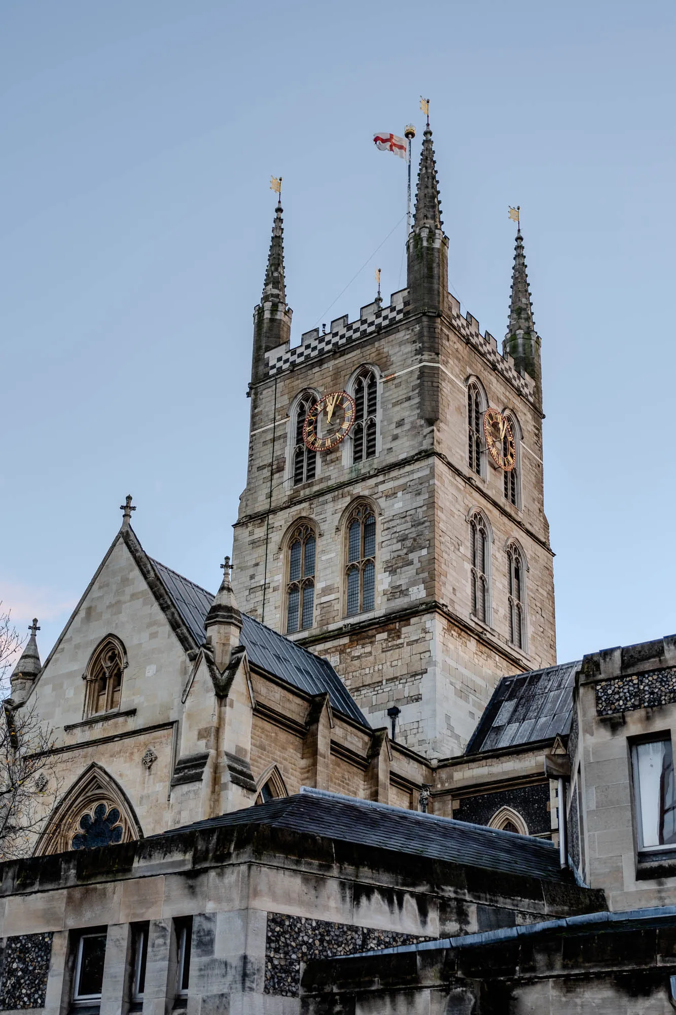 The image shows a tall, stone cathedral with a clock tower.  The tower has a flag flying from the top, which is a white cross on a red background.  The tower has three tall, narrow windows on each side, and a clock face on each of the two sides visible in the photo.  The top of the tower is decorated with a black and white checkered pattern.  To the left of the tower, there is another part of the cathedral with a pitched roof.  This section has a round window at the bottom and a narrow window higher up.  The two sections of the cathedral are connected by a lower, stone building with a black roof.  The sky is clear and blue.