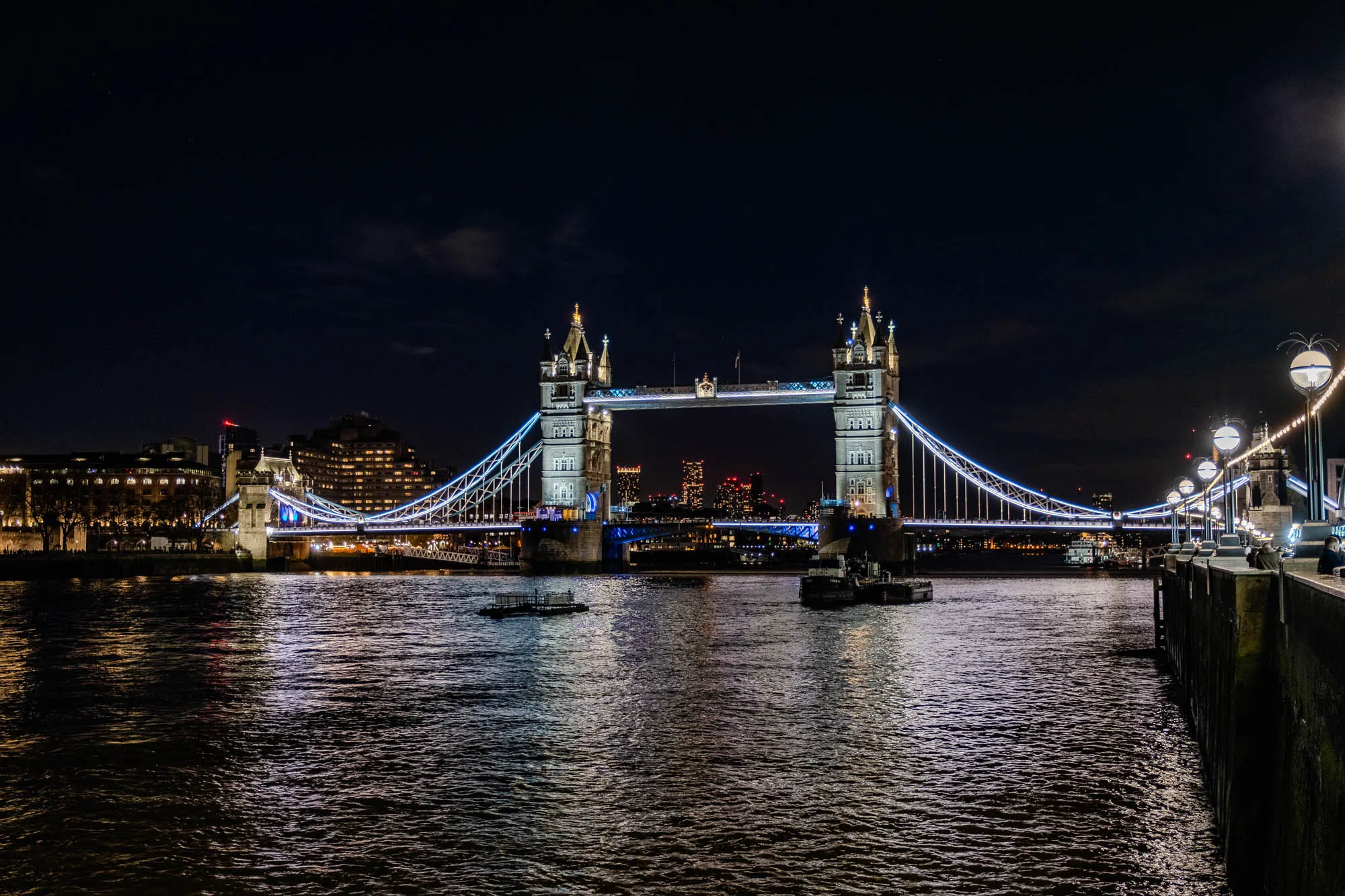 This is a night time picture of Tower Bridge in London. The bridge is lit up with blue lights. The bridge has two tall towers connected by a walkway. The walkway is suspended by cables over a body of water. There are multiple streetlights lining the walkway of the bridge and some buildings in the distance.  The water is reflecting the light of the bridge and the buildings.  The picture was taken from a dock on the right side of the bridge.  There is a small boat on the water in the distance.