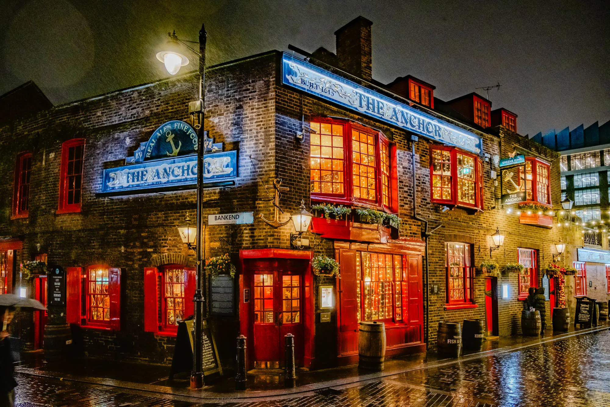 The image is of a pub called "The Anchor" at night, under a light rain. The pub is built of brick and has a large sign above the entrance.  The sign is painted blue and white with the pub's name in large white letters and an anchor design in the center.  The pub's windows are large and have red frames.  The windows are illuminated from inside, and there are Christmas lights strung across the facade. There is a street lamp with a lamppost on the left of the image.  The lamppost has a street sign saying "Bankend" on it.  There are barrels in front of the pub and in front of a side entrance, which appears to be a doorway for people entering.  There is a small awning on the doorway.  There are two people standing in the entrance of the pub, and a person walking on the sidewalk in front of the pub with an umbrella. There is a sign in front of the pub that reads "Real Ales Fine Food" and another sign that says "Greene King."  There is another building visible to the right of the pub. The street in front of the pub is wet with rain.  The rain creates reflections on the cobblestone street.