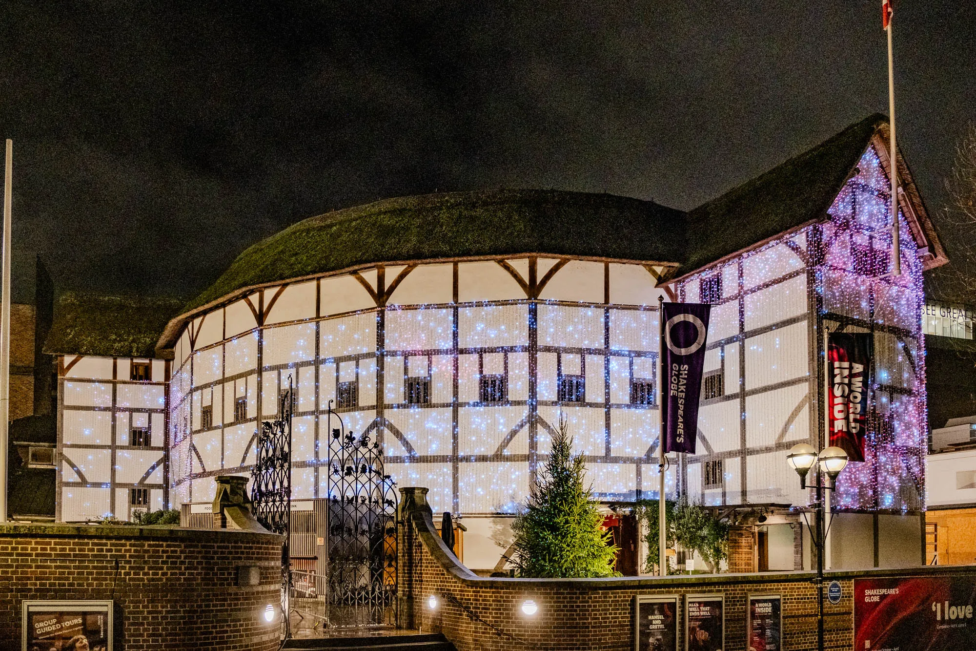 The image shows the exterior of the Globe Theatre in London at night, illuminated by string lights. The building is a large, wooden structure with a thatched roof. It is surrounded by a brick wall and a black metal gate.  The Globe has a sign on the side that reads "Shakespeare's Globe" and another that says "A World Inside."  There is a smaller sign that reads "See Great" on the far right side of the building. The brick wall is covered with posters, including one that reads "Group Guided Tours" and another that reads "'I love." There are a few trees in front of the building and a streetlight on the right side of the image. The sky is dark and the only light comes from the building and streetlights. The building appears to be lit up for the Christmas holiday. 
