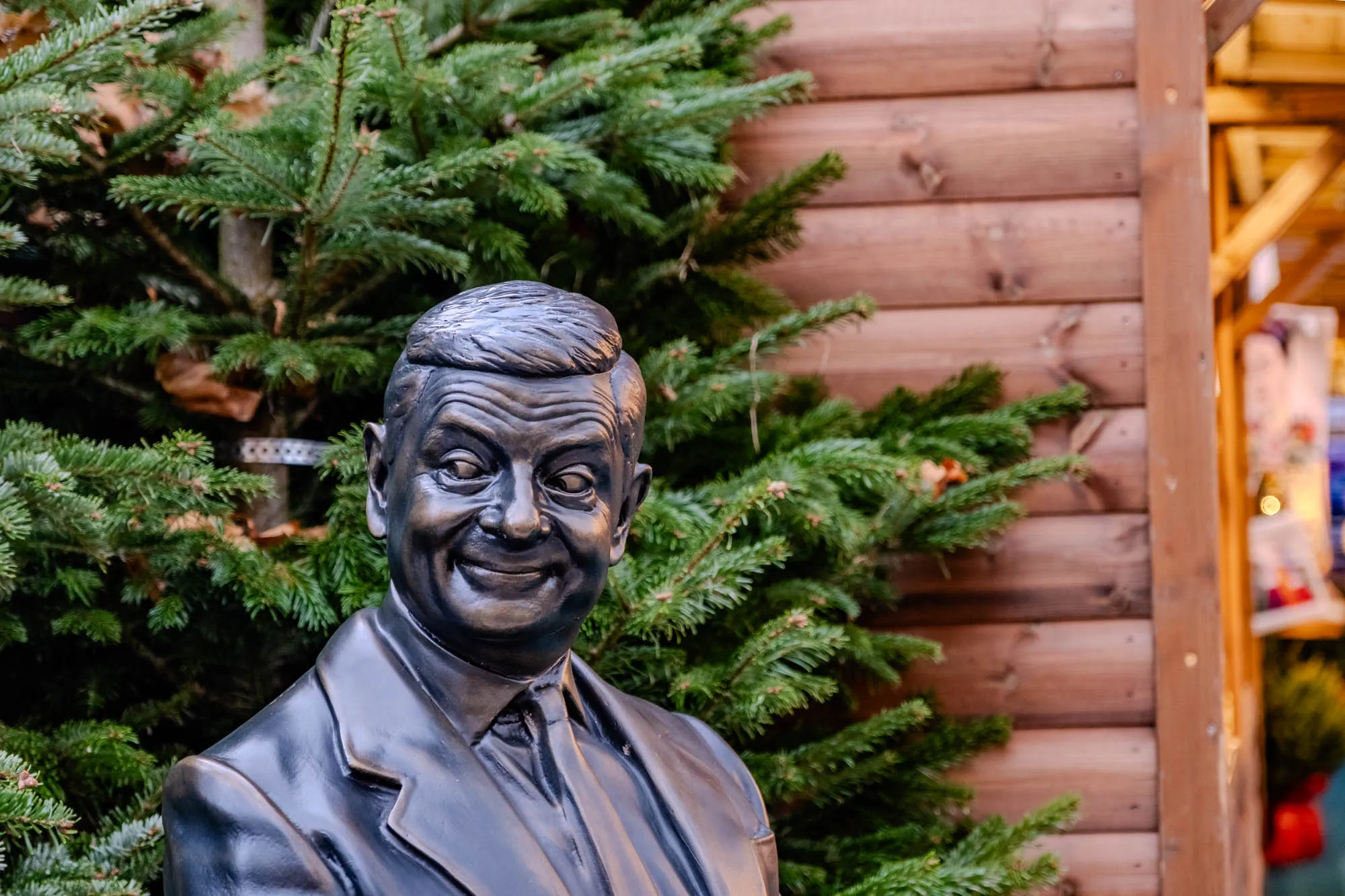 The image shows a bronze bust of a man, Mr. Bean, with a mischievous grin and  wide eyes. The bust is set against a backdrop of a Christmas tree, with its dark green needles and branches partially obscuring the bust. The tree is in front of a wooden building, likely a Christmas market stall, with horizontal wooden planks and a brown vertical post. The building is partially obscured by the tree branches on the left and appears to be lit with warm, yellow Christmas lights.  The image is most likely taken outdoors, during a wintery or holiday season.