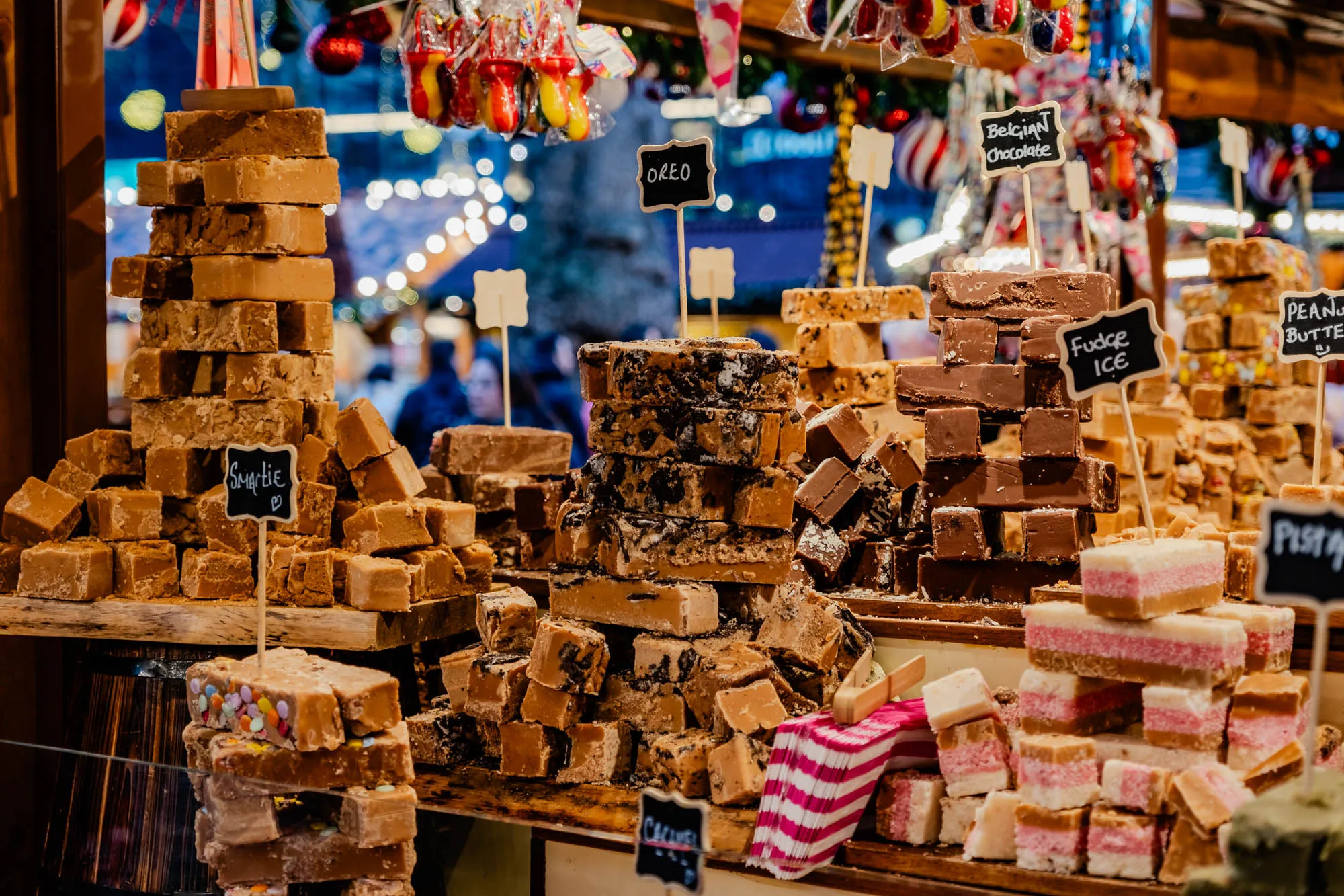 This is a display of fudge, with a variety of flavors.  The fudge is stacked in neat towers behind a glass case.  In the center of the image, there is a tower of fudge labeled "Oreo".  To the right of this is a stack of fudge labeled "Belgian Chocolate".  To the right of this is a stack of fudge labeled "Fudge Ice".  To the right of this is a stack of fudge labeled "Peanut Butter".  To the left of the "Oreo" stack is a stack of fudge labeled "Smartie".  In front of the "Oreo" stack is a display of fudge that looks like it's made with crushed Oreo cookies.  There are also some other flavors of fudge, including some that look like they are made with sprinkles, and some that are pink.  The fudge is arranged in a way that is both appealing and easy to see.