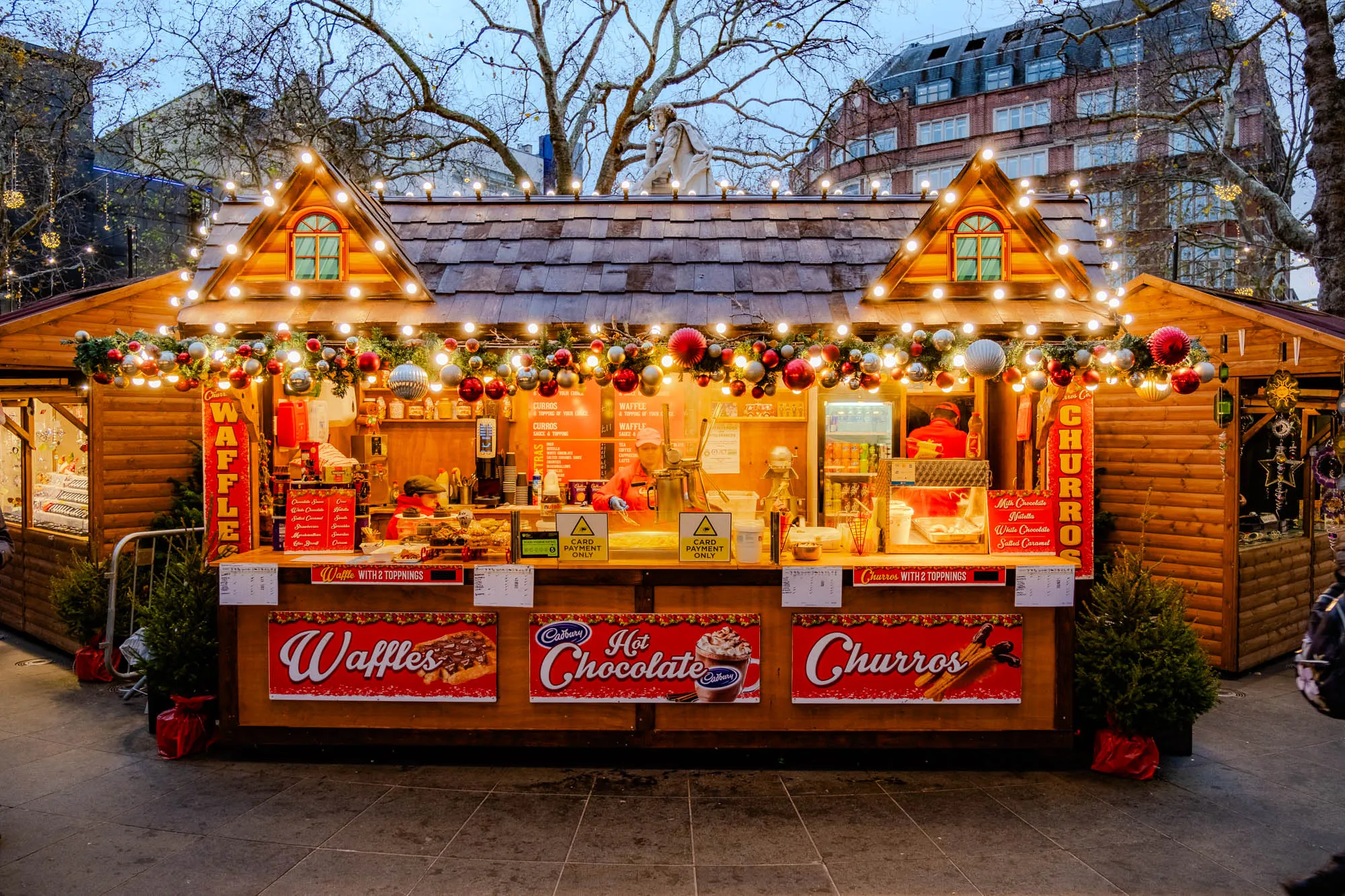 The image shows a wooden food stall at a Christmas market, decorated with Christmas lights and garlands of holly and silver baubles.  The stall is selling waffles, hot chocolate, and churros.  On the left side of the stall is a sign with the word "Waffles" on it, a price list, and an image of a waffle with toppings.  The center of the stall has a sign with "Hot Chocolate" and an image of a cup of hot chocolate topped with whipped cream.  The right side of the stall has a sign with the word "Churros" on it, a price list, and an image of churros.  Behind the stall is a person in a red shirt, and a person standing behind the counter, but their faces are not visible.  There is a small Christmas tree on the ground in front of the stall. The stall is in front of a brick building decorated with Christmas lights.