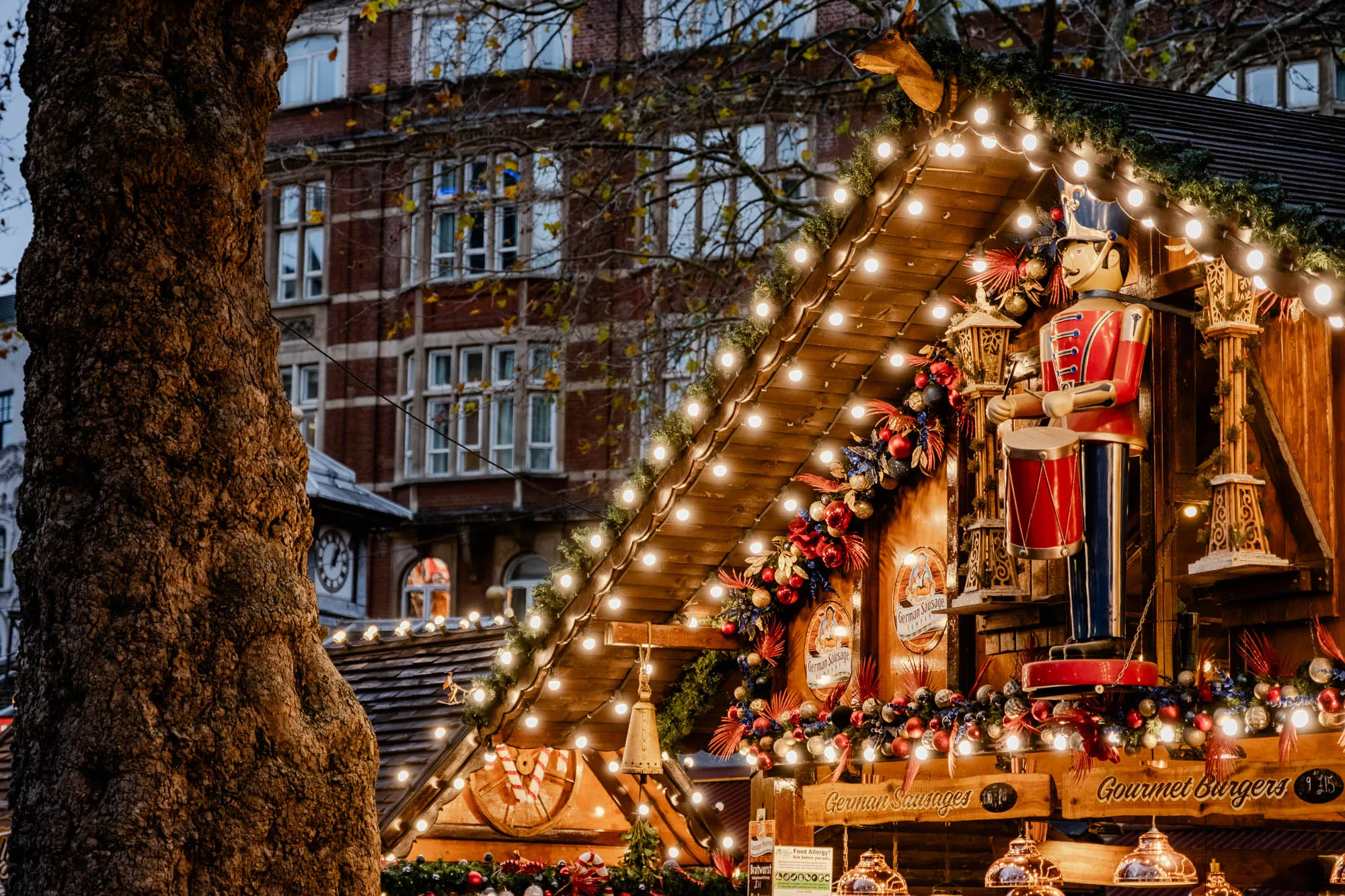 The image shows a close-up of a Christmas market stall. It is a wooden structure with a roof adorned with Christmas lights and garlands of greenery and red and gold ornaments. A large wooden nutcracker doll stands on a platform in front of the stall, holding a drum. The nutcracker is dressed in a red and blue uniform. The stall is lit by warm white lights that cast a soft glow over the scene.  The stall is labeled “German Sausages” and “Gourmet Burgers”. The sign is a wooden plank with a large handwritten font. Behind the stall, we see a tall brick building, likely an apartment complex, with multiple windows and a tree in the foreground on the left side. The building is partially obscured by the tree, and the focus is on the Christmas market stall.  The image is a festive and cheerful scene that evokes the magic of the holiday season.