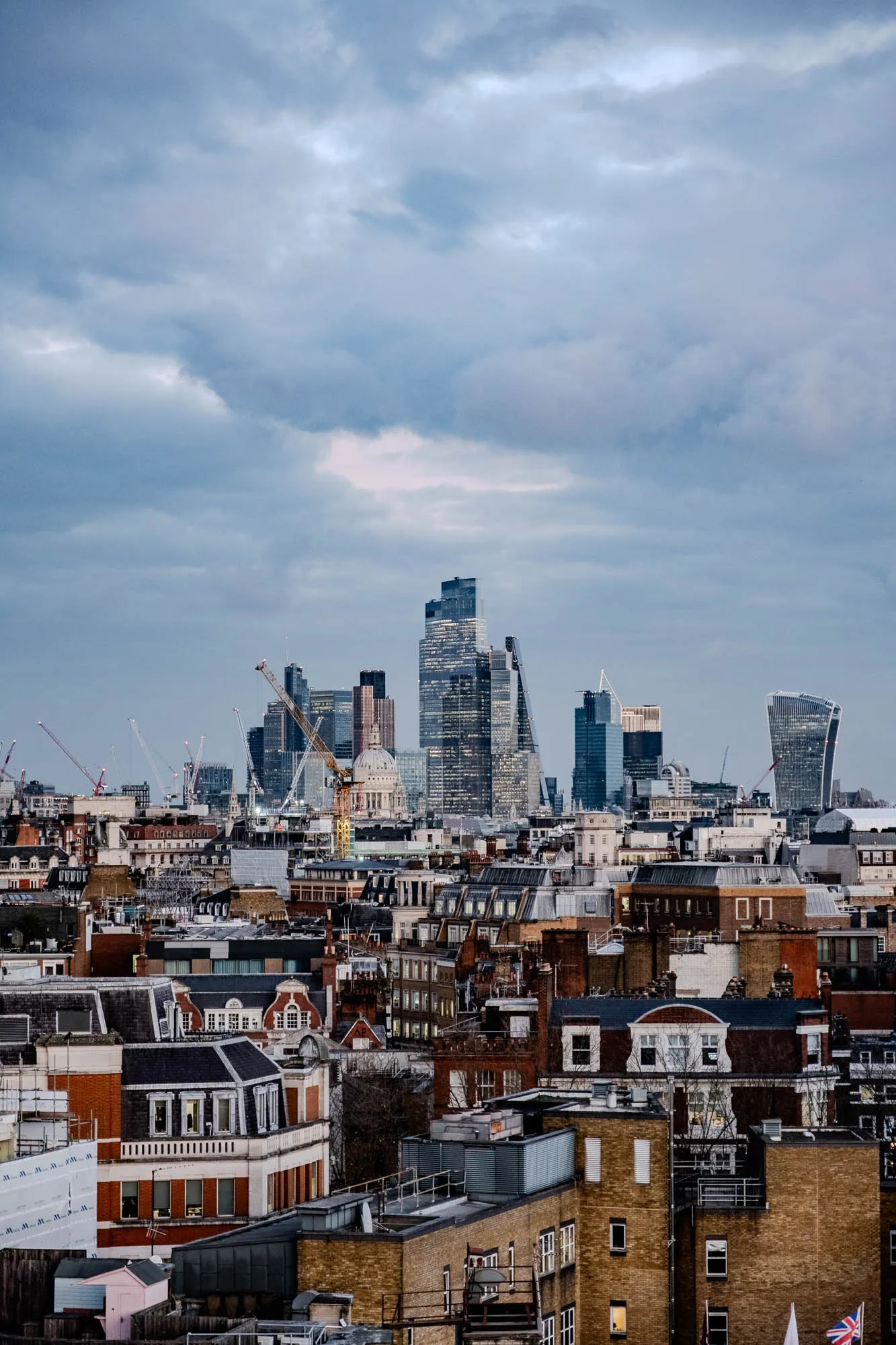 A view of a city skyline with a cloudy sky. There are many skyscrapers in the distance, including one that is very tall and thin. There are also many smaller buildings in the foreground. There is a construction crane in the center of the image.