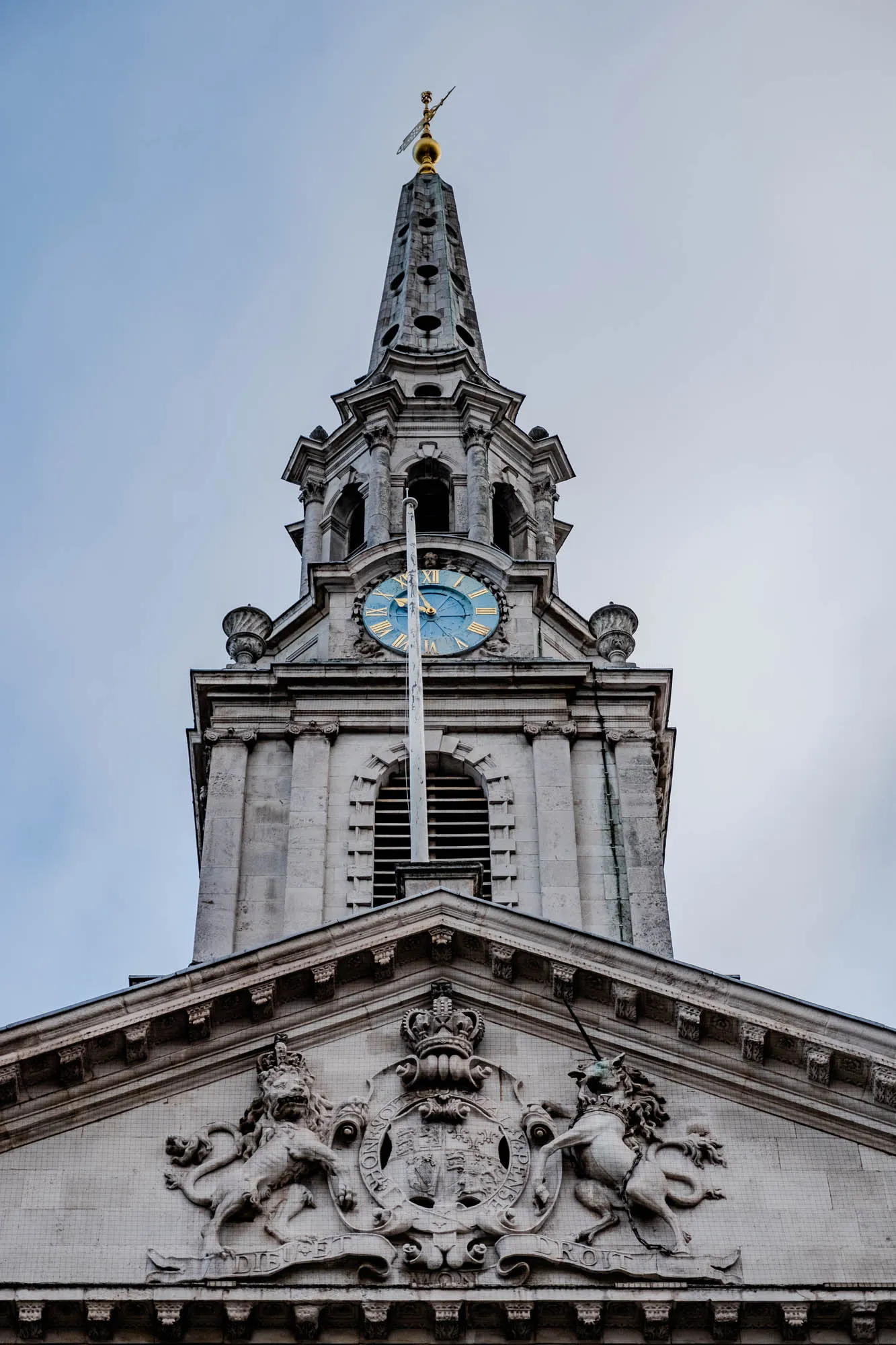 The image is a low angle view of the top of a church with a tall spire.  The building is made of stone and has a clock face on the front. The clock face has Roman numerals and a blue face with gold hands. Above the clock face are several arches and decorative elements, including a spire topped with a weather vane. The spire is topped with a golden ball. The building is very ornate with a lot of decorative elements.  Just below the clock face are carvings of a lion and a unicorn. The lion is on the left side and the unicorn is on the right side.  The lion and unicorn are both facing toward the viewer. They are both standing on a scroll with an inscription that reads, "Dieu et Mon Droit". There is a coat of arms between the lion and unicorn. The coat of arms is also very ornate, and it features a crown, a lion, and a unicorn.  The background is a light blue sky.