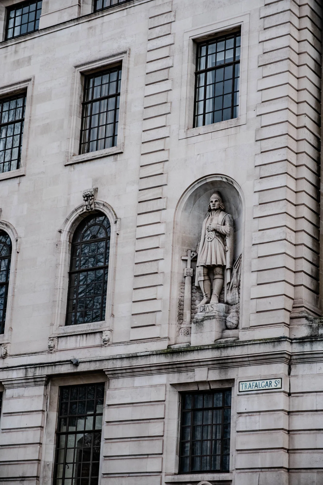 The image is of a building's facade, which is made of white stone and has a lot of ornate details. There are multiple windows, including one large arched window on the left side of the image. The building also has a stone statue of a man in a niche in the wall. The statue is of a man with a beard and mustache wearing a long robe and holding a sword. The statue is labeled “Dias 1488” in the niche. At the bottom right of the image, there's a sign that reads “Trafalgar Sq.”