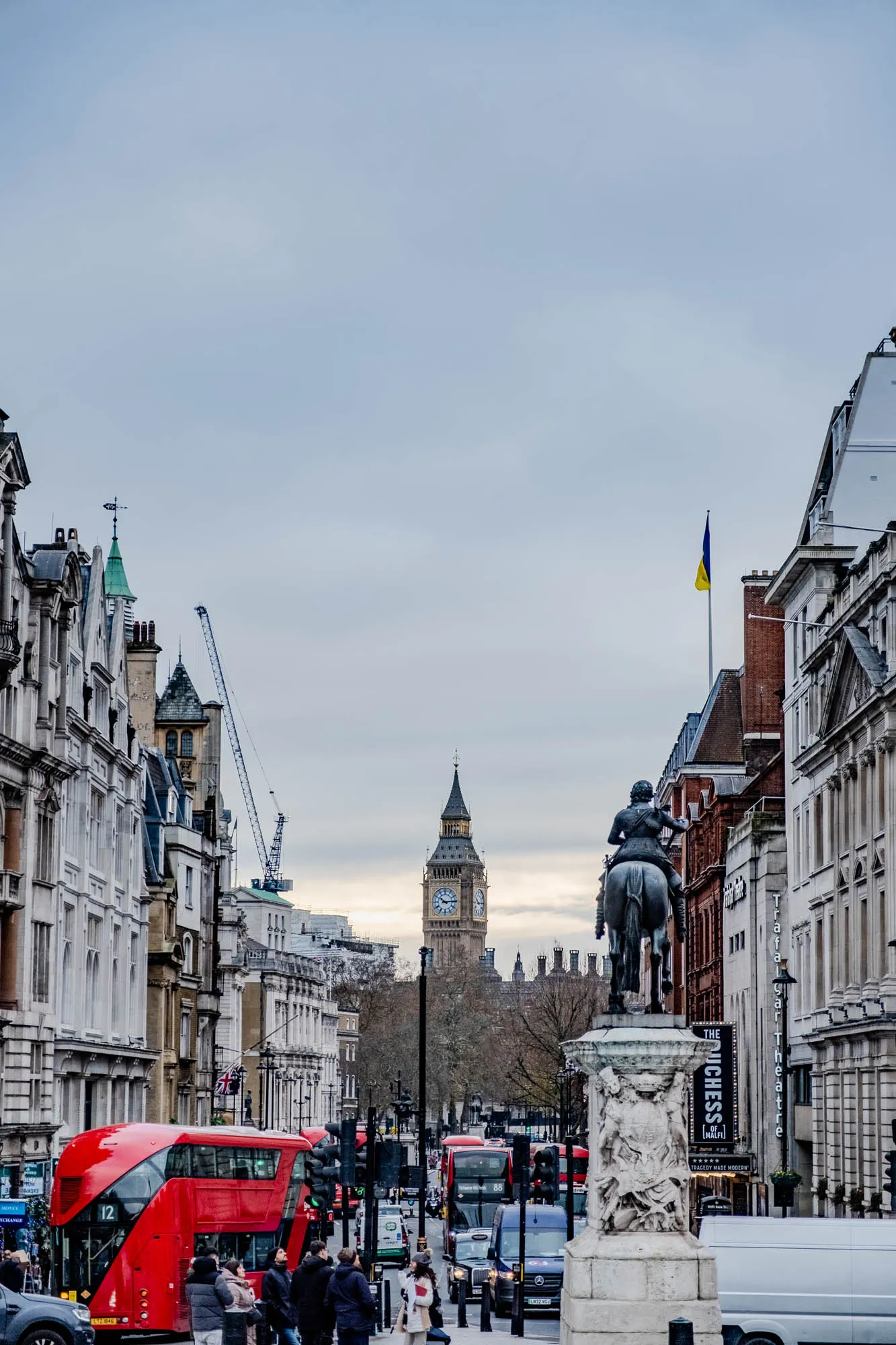 The image shows a street in London, England. The street is lined with buildings on both sides, many of which are tall and old. In the distance, you can see Big Ben, a famous clock tower.  A statue of a man on horseback sits in the middle of the street, and the street is busy with people and cars. The street is lined with traffic lights and traffic signs. There is a yellow and blue flag on a pole on the right side of the picture. To the left of the picture, there is a double decker red bus. The sky is overcast and gray.