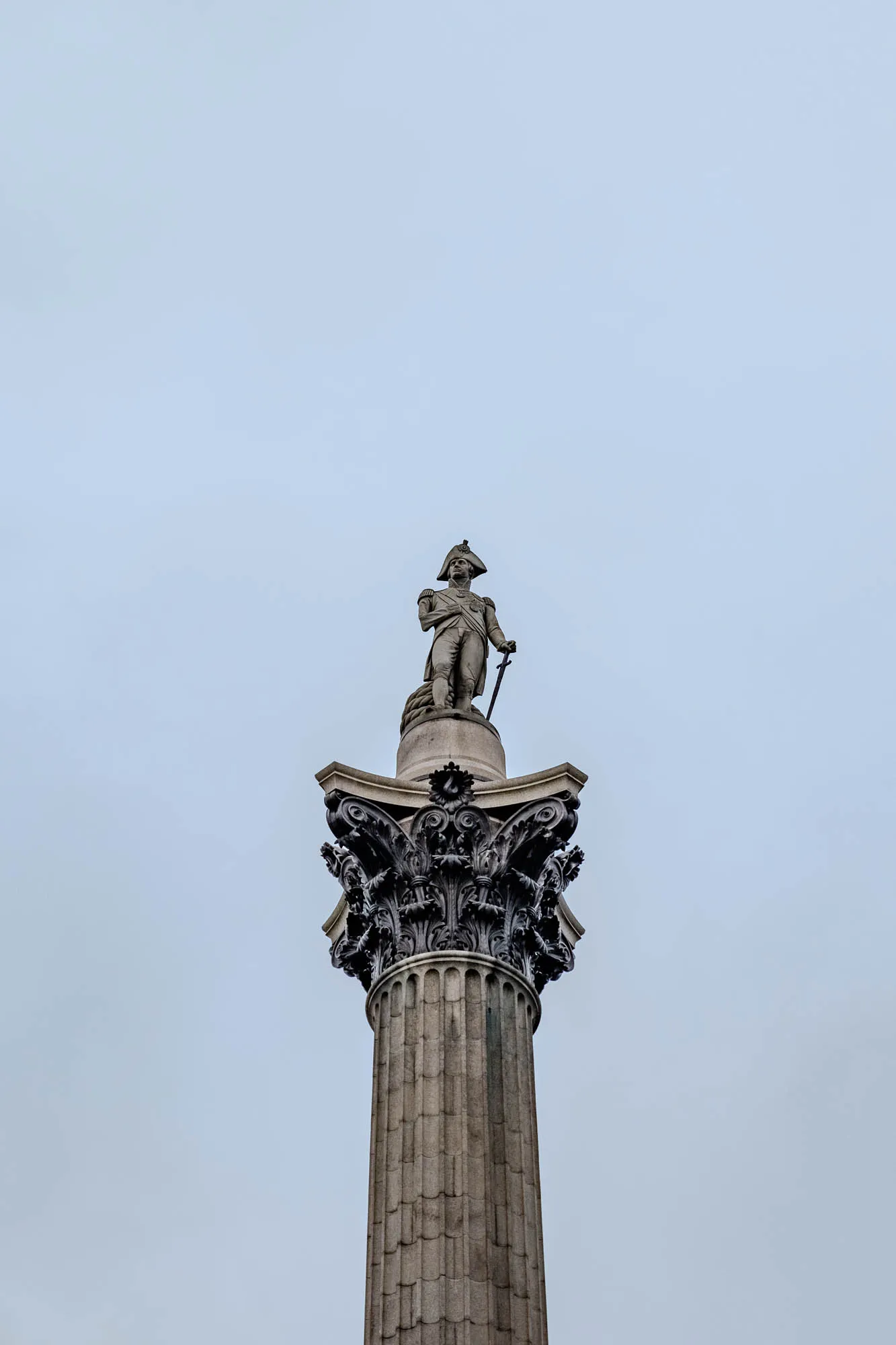 The image shows a tall, grey stone column topped with a statue of a man in a military uniform, holding a sword. The man has a hat on his head. The column has decorative detail around its base. The background is a pale blue sky.