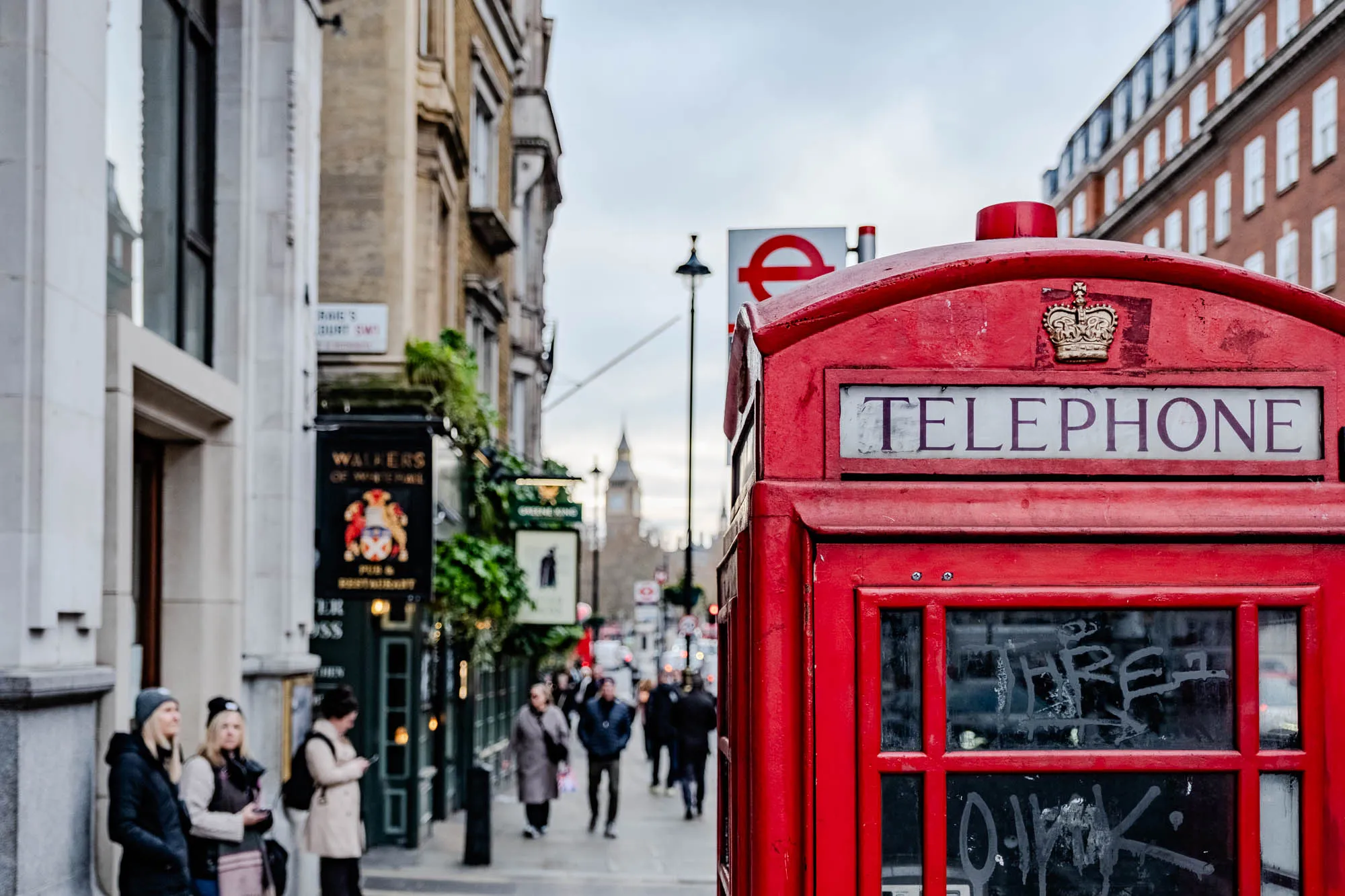 The image shows a class red London telephone booth on a city street. The booth is brightly lit and has a sign above it that reads "TELEPHONE". The booth is slightly angled towards the viewer, so the right side of the booth is more prominent. The front window of the booth has been defaced with graffiti. There is a small gold crown on the top of the booth. In the background, there are pedestrians walking along the street. The buildings on either side of the street are tall and gray. The sky is cloudy and gray. Big Ben is out of focus in the background.