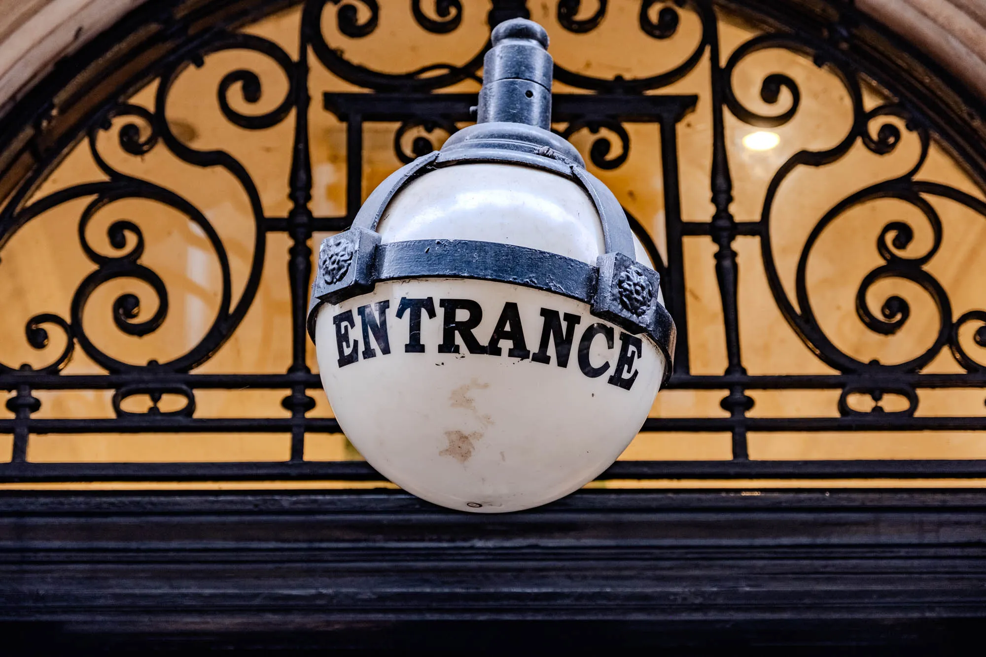 The image shows a close-up of an old-fashioned, round, white light fixture that is hanging from the ceiling of an entrance. The light fixture is made of black metal and has a black metal band with the word “ENTRANCE” written in large, bold, black letters on the front of it. Behind the light, surrounding it, is a black, decorative, metal lattice in front of window. The lattice has ornate, swirling designs. The window is made of glass with a frosted or translucent finish. The light is off.  Behind the lattice, the ceiling is a solid, light, brownish yellow color.  The top and bottom of the image is a dark, black, wooden molding that surrounds the lattice and light fixture.