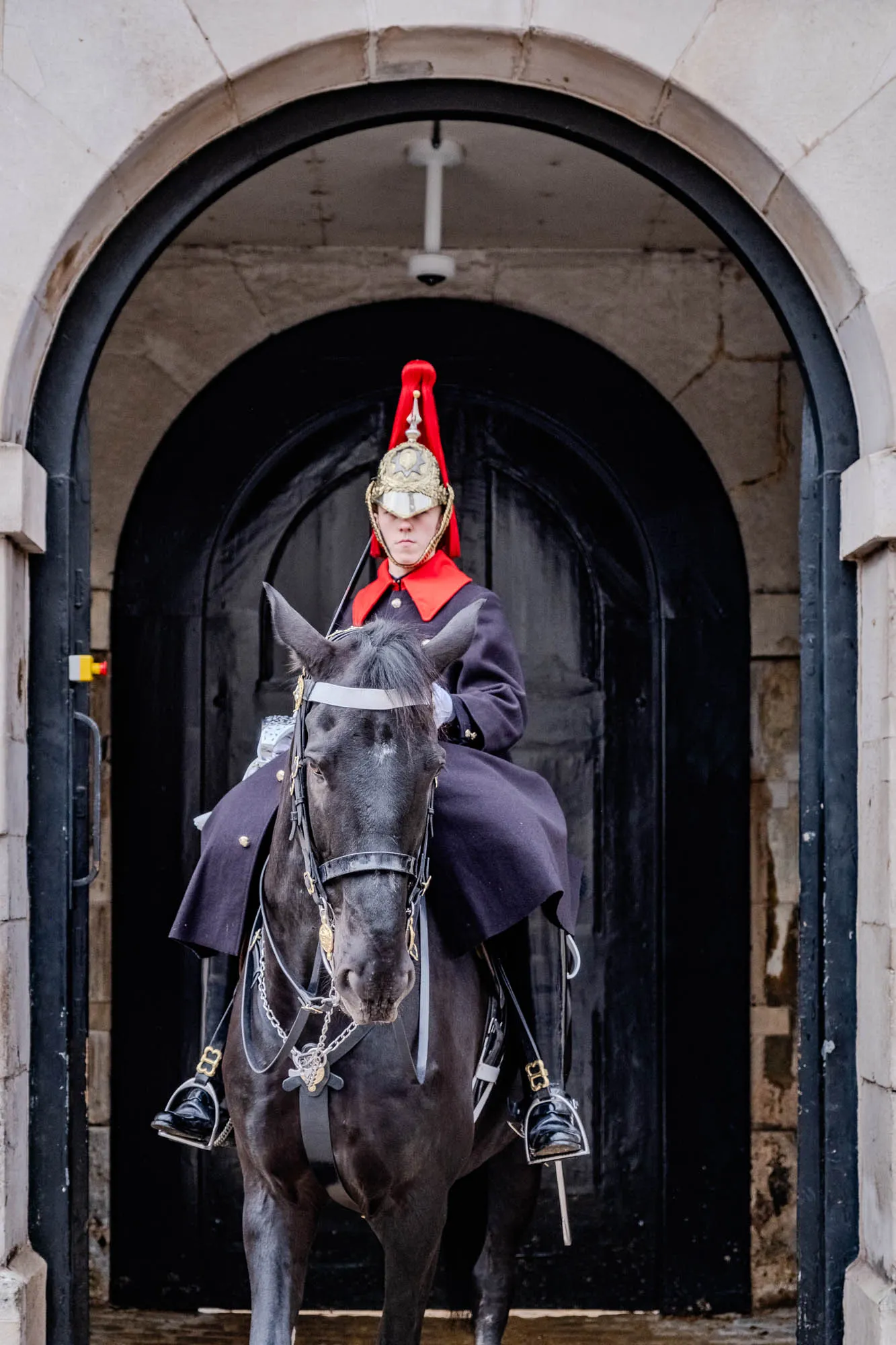 The image shows a person on horseback. The person is wearing a black uniform with a red and gold helmet. They are riding a black horse with a white strap across its forehead. The horse is wearing a leather bridle with a silver chain attached. They are standing in front of an archway made of stone with a black door. The person is looking straight ahead.  The background is out of focus.