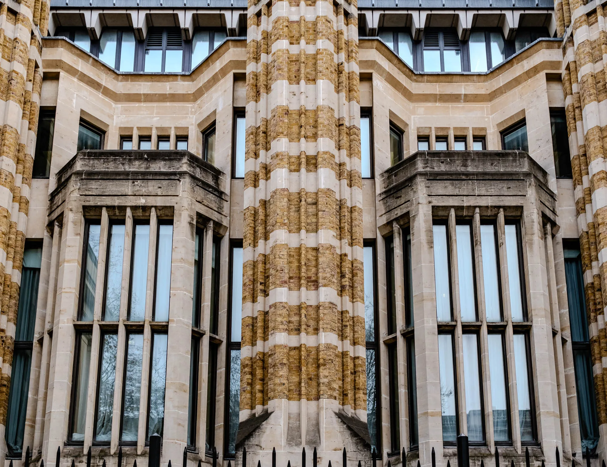 This is a photo of a building. The building is made of light colored stone with dark brown trim around the windows. The windows are all rectangular and have multiple panes. In the middle, there is a column made of tan brick. The brickwork in the column is striped, with bands of white brick alternating with bands of brown brick. The photo is taken at a low angle, looking up at the front of the building. You can see a black metal fence in the foreground, which is partially obscured by the building. The building has a symmetrical design, with the left side of the photo looking like a mirror image of the right side.