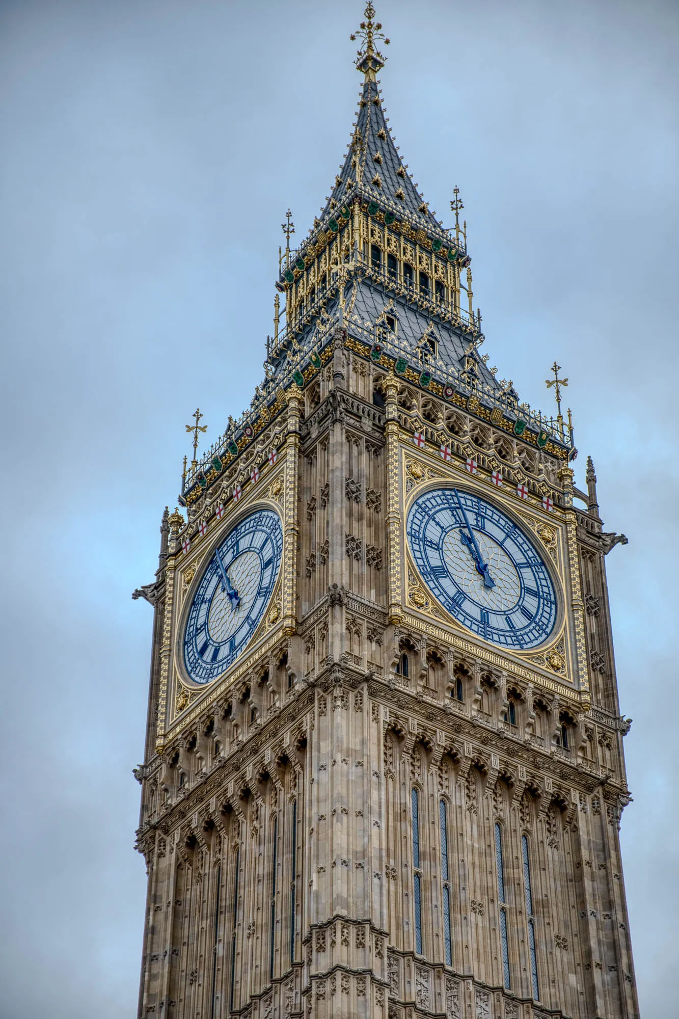 The image shows the iconic Elizabeth Tower, also known as Big Ben, in London. The tower is a tall, stone structure with a pointed roof. It has a large clock face with Roman numerals and black hands. The clock face is surrounded by a gold frame. The tower has many intricate details and is made of stone and metal. The sky is cloudy. The tower is a symbol of London and the United Kingdom.