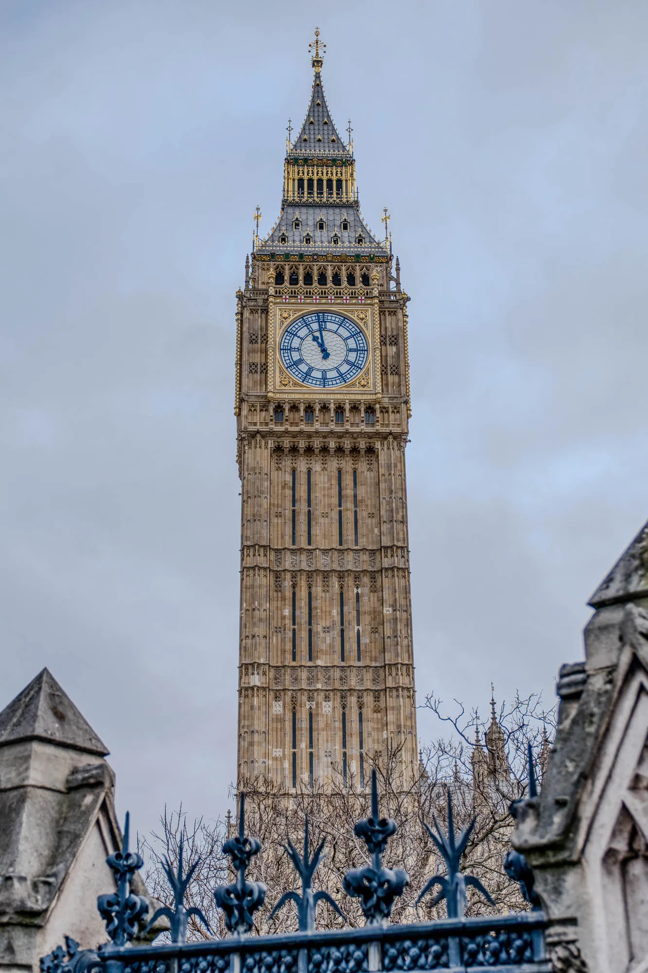 The image shows Big Ben, the famous clock tower in London, England. The tower is tall and slender, with a pointed roof. It has a large clock face with Roman numerals, and the hands are pointing to the time. The tower is made of stone, and it is decorated with intricate carvings. The tower is seen through a wrought iron fence with a grey stone structure on the left and right of the image. The sky is overcast and there are bare branches of trees in front of the tower.