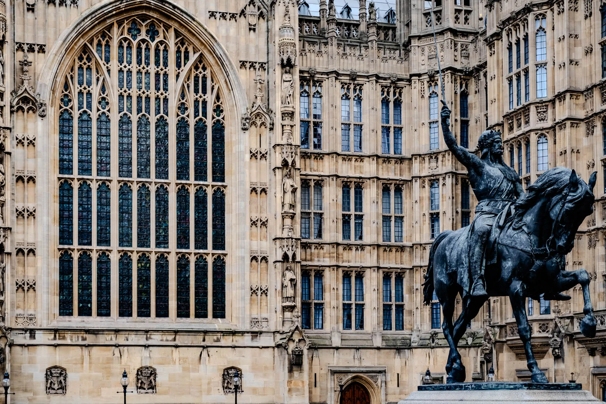 The image shows a statue of a man on horseback in front of a large stone building. The man is holding a sword in his right hand and is wearing armor. The horse is also armored. The building has many windows and arches. The stonework is detailed and intricate. The image is taken from a low angle, so the statue appears large and imposing. There are streetlights on either side of the statue. There are two decorative coats of arms or plaques on the wall of the building, below the large arched window.