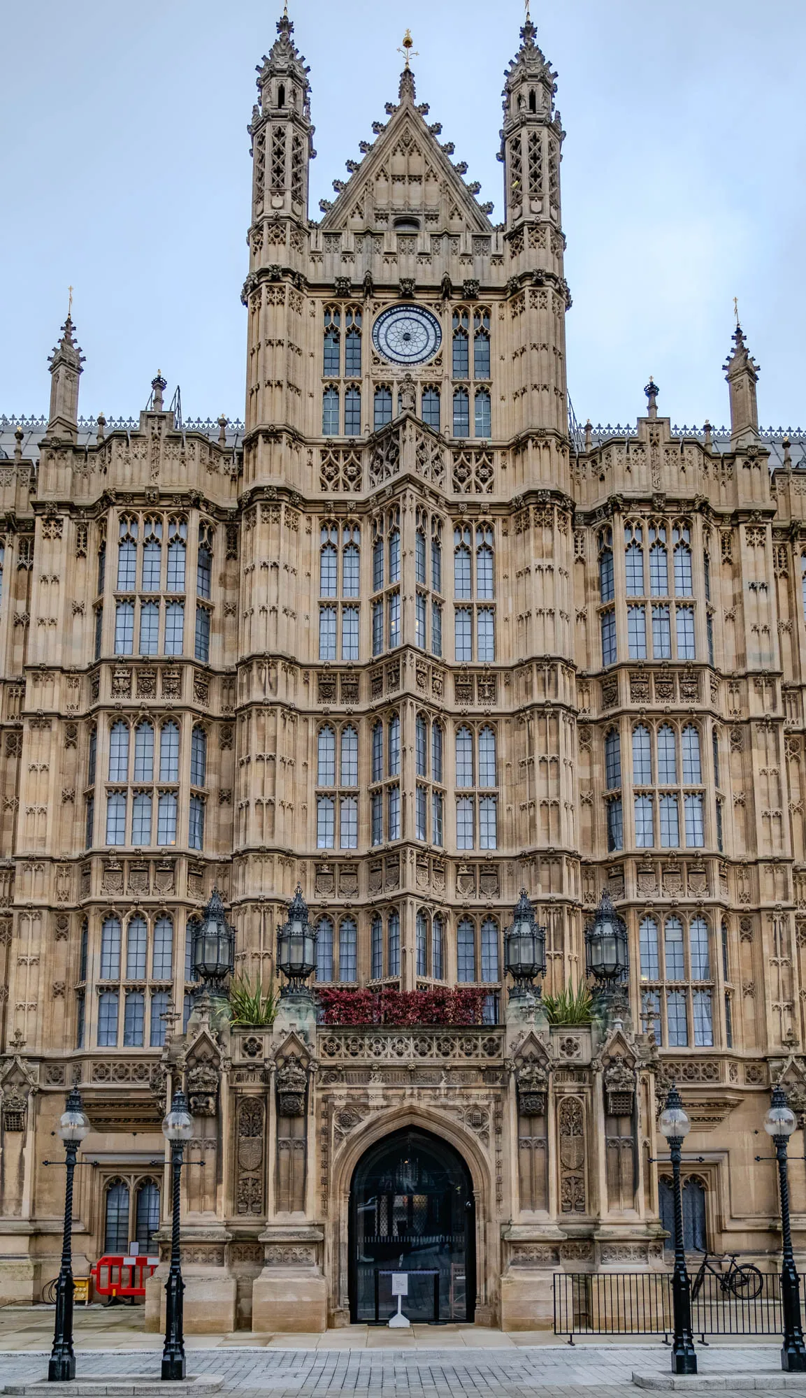 The image shows the front of a large, ornate building. The building has two tall, pointed towers on either side of the center. There are many windows with arched tops. The building is made of stone and appears to be very old. In the center of the building is a large archway leading to a door. There are black lampposts on either side of the archway and a metal fence in front of the building.  The ground in front of the building is a paved walkway with grey square tiles.  There are two bikes in front of the building.  A red barrier and yellow construction signs are visible on the left side of the building.  The building is likely a government building.