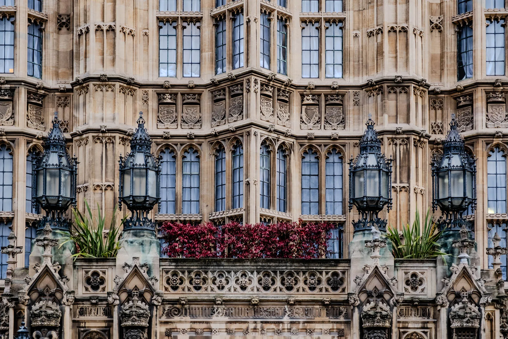 The image shows a close-up of the facade of a large, ornate building. The building is made of stone and has a lot of intricate detail. There are many windows, some of which are covered with curtains. Above the windows are several coats of arms. There are two large, ornate lampposts in front of the building. Between the lampposts is a small balcony with red plants.  There are also many sculptures of faces and other figures on the building.