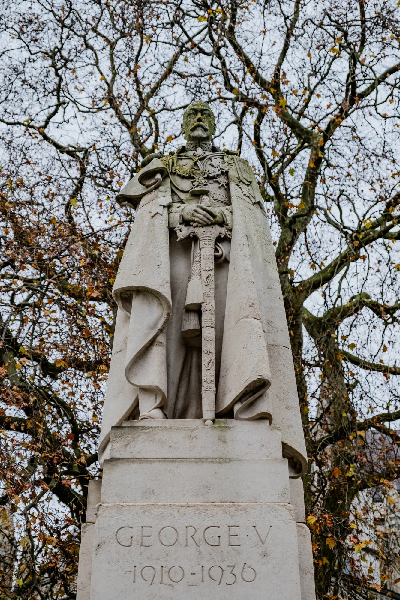 The image shows a stone statue of a man wearing a long coat and a sash, standing in front of a tree with bare branches and some yellow leaves.  The man is holding a sword in his right hand and his left hand is resting on the hilt. The statue is on a tall stone base with the words "GEORGE V" and "1910-1936" inscribed on it. The statue is in the middle of the image, with a blurred the tree behind it and a blurred building to the bottom right.