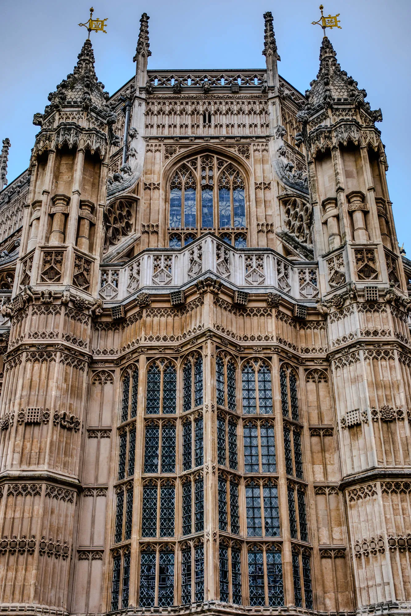 The image shows a close-up of the exterior of a large, ancient building. The building is made of stone and has a lot of intricate details, including tall, pointed arches, decorative carvings, and a series of tall, narrow windows.  The windows are divided into smaller panes by thin, dark mullions.  The stonework is a light brown color and appears to be quite weathered. The building is likely a church or cathedral, given its architectural style. There is a clear blue sky in the background, making the building appear even more majestic. The top of the building is capped by two spires, which are topped with gold ornaments. The whole image suggests a building of great age and historical significance.