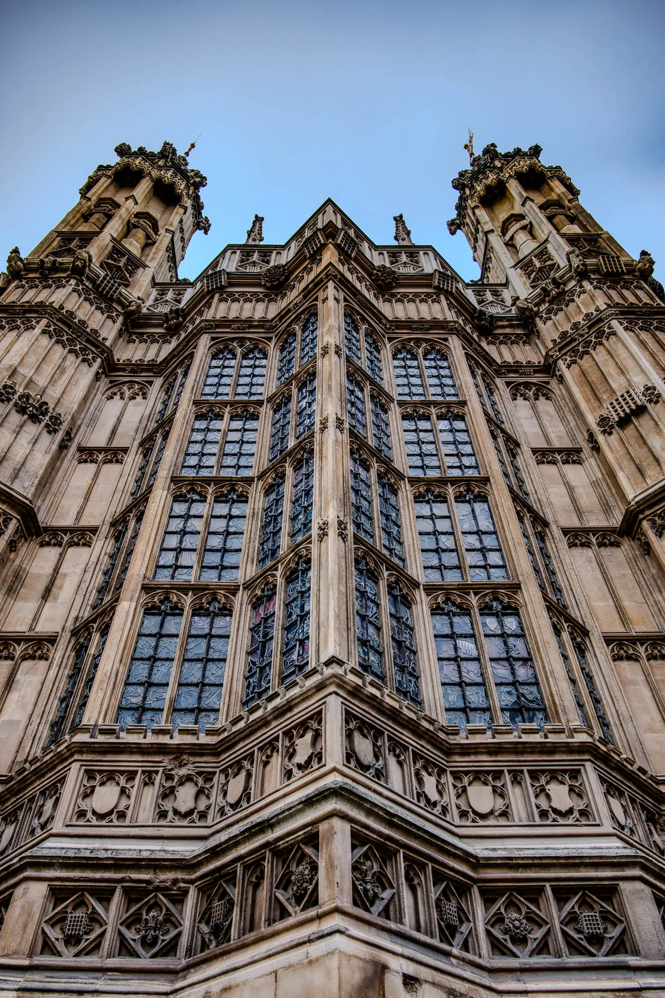 The image is a low angle shot of a stone building. The building appears to be a church or cathedral, with a large stained glass window in the center of the image. The window is made up of many smaller panes of glass, and there are carvings around the edge of the window. The building is made of stone, and there is a lot of intricate detail in the carvings. The image is taken from a low angle, so the building appears to be very tall. The sky is blue and there are clouds in the distance.  The image is taken from a low angle, so the building appears to be very tall. The sky is blue and there are clouds in the distance.  The building has two towers on either side of the window.  The towers have a lot of intricate detail in the carvings.  There are carvings on the exterior walls of the building that are below the window. The image is a good example of the architectural style of the building.