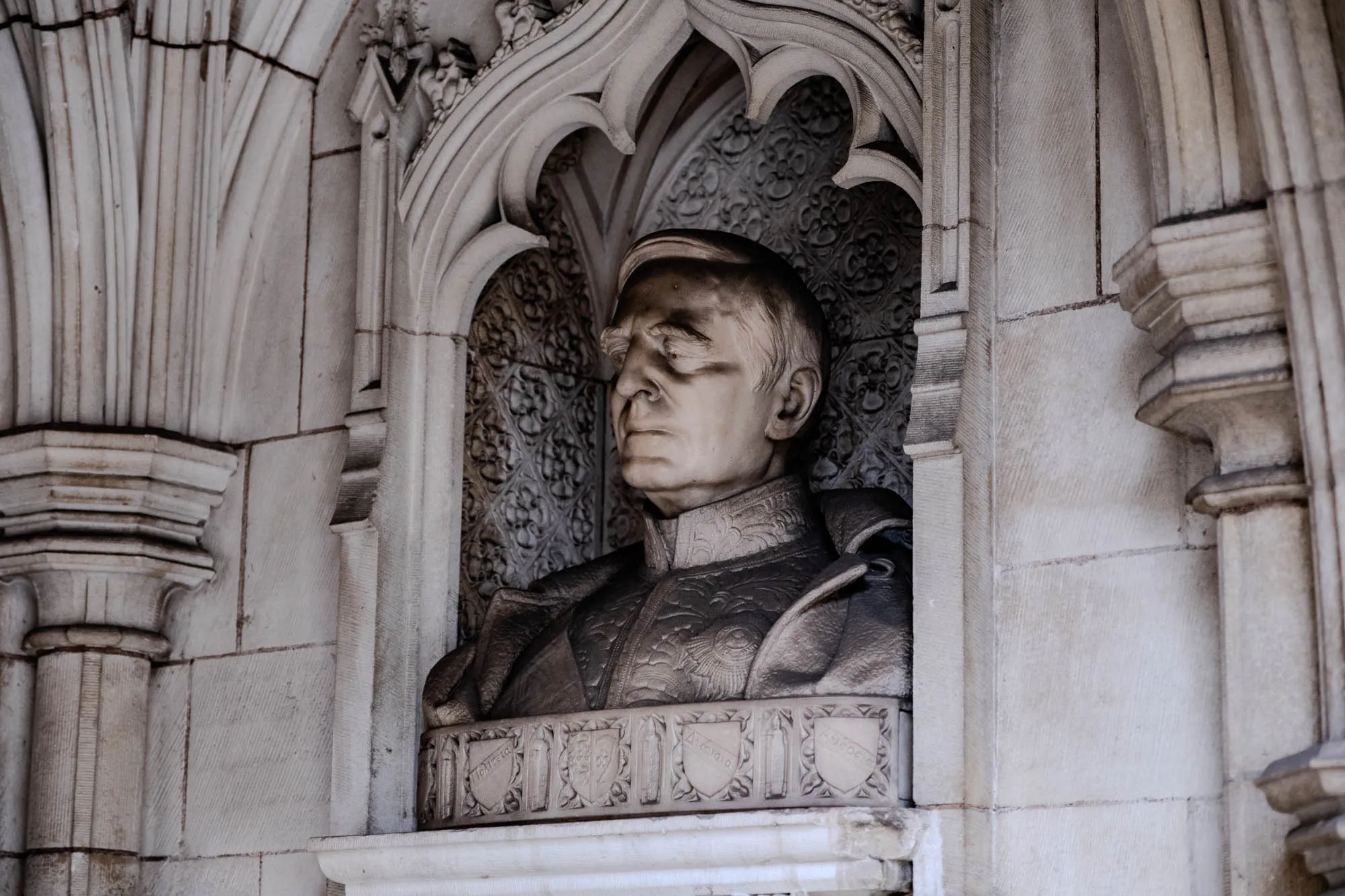 The image shows a stone bust of a man set into a stone wall. The bust is of a man with short, gray hair and a mustache. He is wearing a jacket with intricate detailing. The bust is set into a niche in the wall. The wall is made of large, rectangular stones. The wall has a decorative archway around the bust and a series of ornate, carved shields below the bust. The image is in black and white.