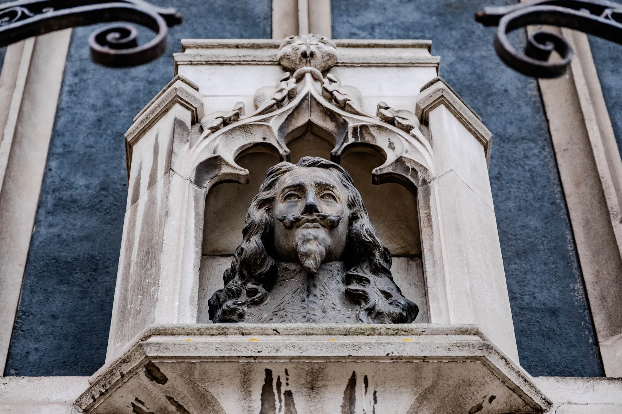 A stone bust of a man with long hair and a mustache is set in a niche. The niche is part of a stone building, and the man's head is just below a stone archway. The man has a serious expression on his face. There is a wrought iron railing in the background and the wall is a dark grey.