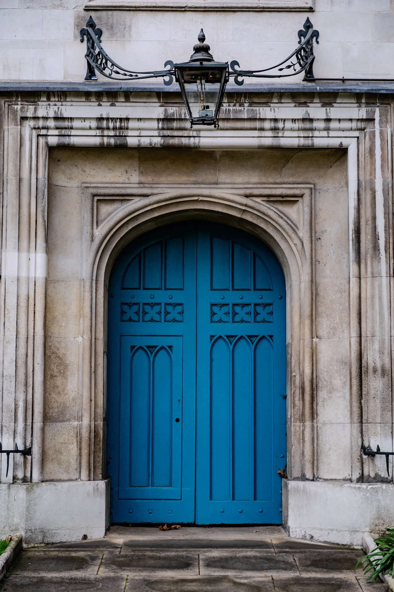 The image shows a large, ornate, blue door with two panels. The door is set within a stone archway, with an old-fashioned street lamp mounted above it. The door is made of wood, and has a decorative pattern carved into the panels. The door is set within a stone archway, and there is a stone ledge running along the bottom of the arch. The door appears to be a part of a stone building, and there is a stone walkway leading up to the door.  The stonework has signs of weathering and aging. There are two small iron railings to either side of the doorway.  To the left, just beyond the railing, there is a small patch of grass. To the right, there is a small patch of grass growing in a flower bed.  The scene looks like it could be in a European city.