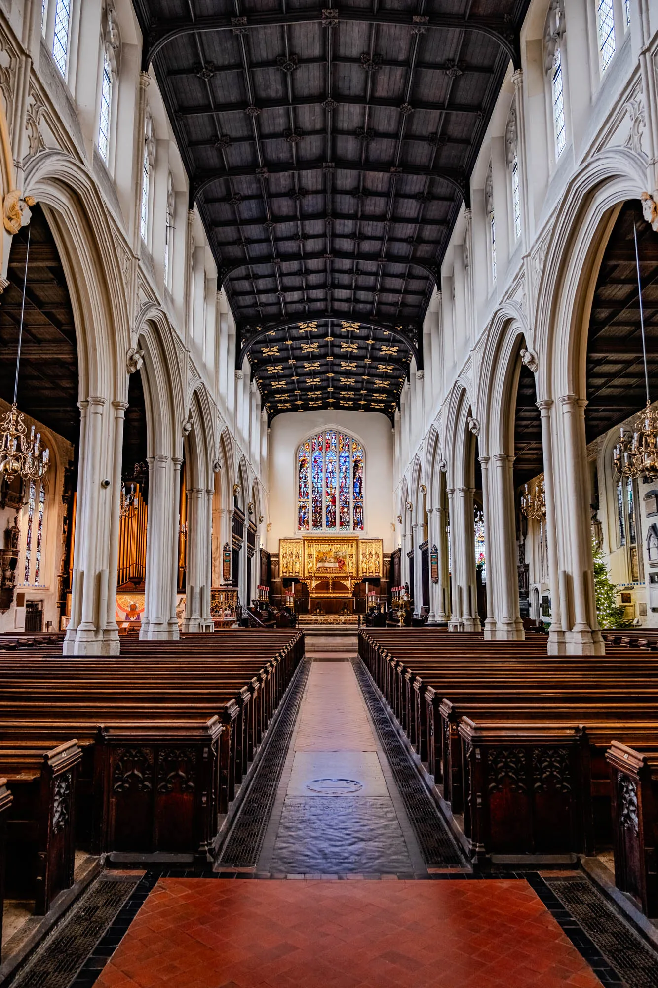 The image shows the interior of a church. It has a long, central aisle with rows of wooden pews on each side. The aisle is lined with a dark, grey patterned stone floor. The ceiling is high and wooden with a pattern of squares and decorative carvings. The walls are painted white and have tall, arched windows. There is a stained glass window at the far end of the aisle, which is filled with colorful glass and depicts a scene of a biblical story. The window is framed by a golden archway with a golden altar behind it. There is also a pipe organ on the left side of the aisle. The church is lit by chandeliers hanging from the ceiling. It is a place of worship and prayer.