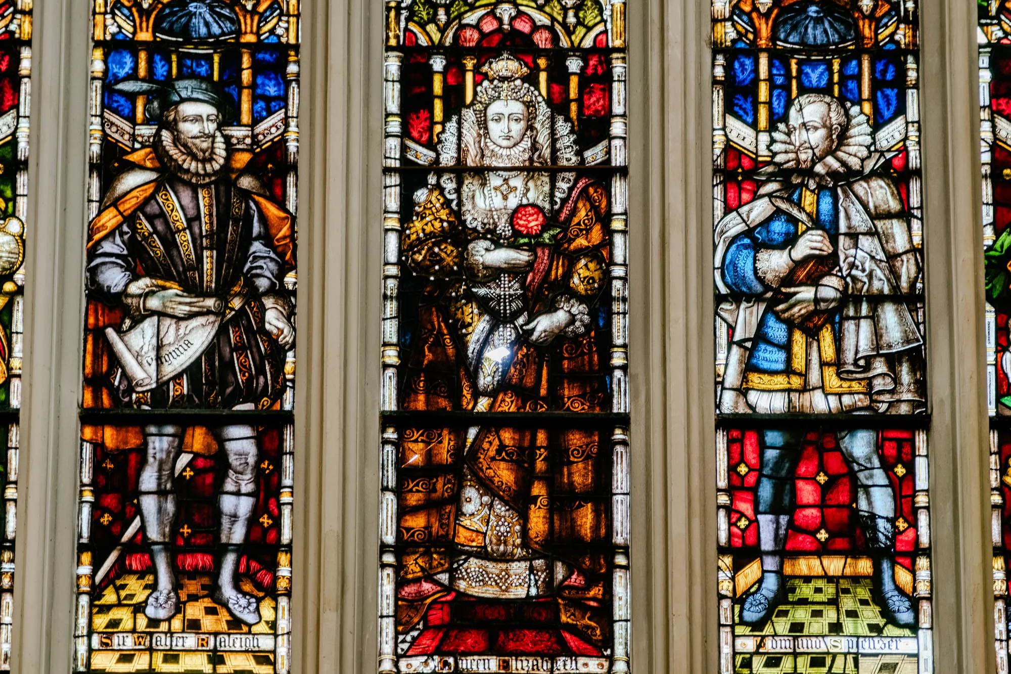 This is a stained glass window with three sections. The middle section has a woman in a long gown and a crown on her head. The woman is holding a red rose in her hand. She has a pearl necklace on and is wearing a white ruff around her neck. 

On the left side of the woman is a man wearing a black hat, a dark jacket, and a large ruffle collar. He is holding a scroll in his hands that says "Virginia" on it. The man has a dark beard and is wearing a gold belt.

The right side of the woman has a man in a blue jacket and a white ruff around his neck. He is holding a scroll in his hand and has a light beard. The man is wearing a red cloak and is standing on top of a patterned rug.

All three individuals are framed with elaborate gold, red, and blue stained glass designs. The bottom of each section has a line of text. The left section reads "Sir Walter Raleigh" and the right section reads "Edmund Spenser", and the middle section reads "Queen Elizabeth".