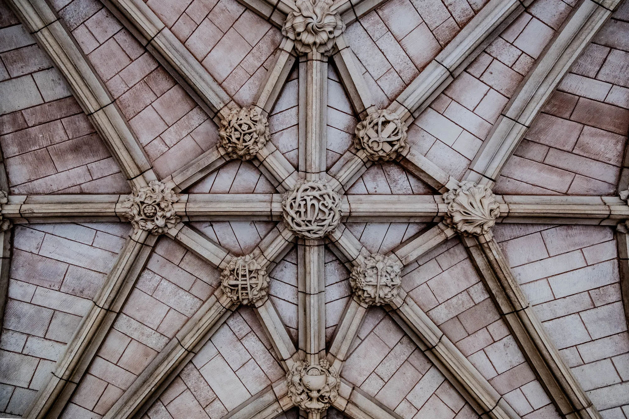 The image shows a vaulted ceiling composed of white stone bricks, with a pattern of intersecting arches that meet in the center.  There are eight stone ornaments placed at the intersections of the arches.  The central ornament appears to have the letters IHS carved into it.  The other ornaments are a variety of carvings, including leaves, flowers, and other designs.  The image gives a sense of the grandeur and intricate detail of a church ceiling.