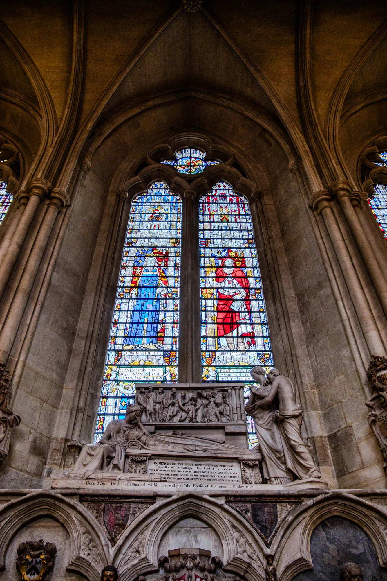 This image depicts a memorial in a stone cathedral. In the center of the image is a large stained glass window. The window has three sections with ornate designs. The top section contains a small circular design. The two lower sections are more rectangular and feature detailed patterns and figures in reds, blues, and yellows.  Under the window is an elaborate stone memorial with a small inscription on a plaque. The inscription says "In memory of the right Hon' Spencer Perceval, Chancellor of the Exchequer, First Lord of the Treasury. This monument was erected by the Prince Regent and Parliament to record their deep sense of his public and private virtues and the Nation's abhorrence of the act by which he fell." Under this, the date of his birth and death are listed as "Born 1 Nov. 1762. Assassinated within the walls of the House of Commons, 11 May 1812." On either side of the main inscription are more elaborate stone carvings and the remains of an ornate stone coffin with the figure of a man and woman sitting on the top.  The cathedral's high vaulted ceilings are visible above the memorial, and the overall tone is somber and reverent.