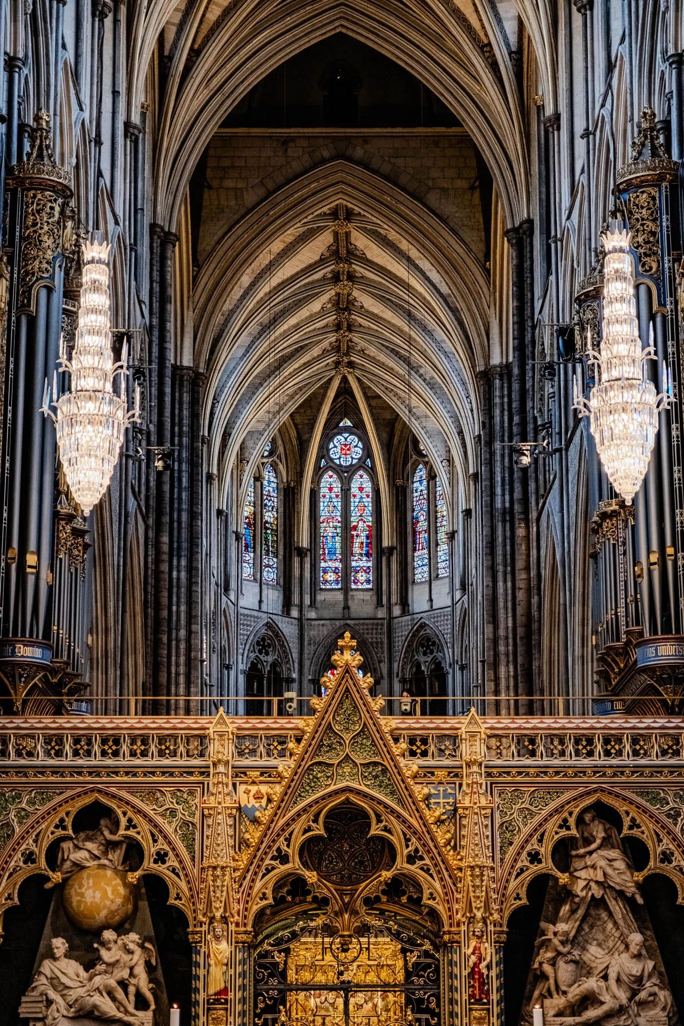 The image shows the interior of a large cathedral. The ceiling is very high and is made of stone arches with intricate carvings and gold accents. The walls are made of stone and are covered with arches. There are two large stained glass windows in the center of the image. The windows are made of colorful glass panels and depict scenes from the Bible. There are two large chandeliers hanging from the ceiling, each with dozens of crystal prisms. The chandeliers are reflecting light from the windows.  The lower part of the image shows the intricate carvings and gold accents of the altar area, with a partially obscured golden shrine in the center. The ornate decorations are primarily gold, with some green and blue accents. There are two stone sculptures flanking the shrine. The sculptures are of people kneeling in prayer. One sculpture is a male and the other is a female.