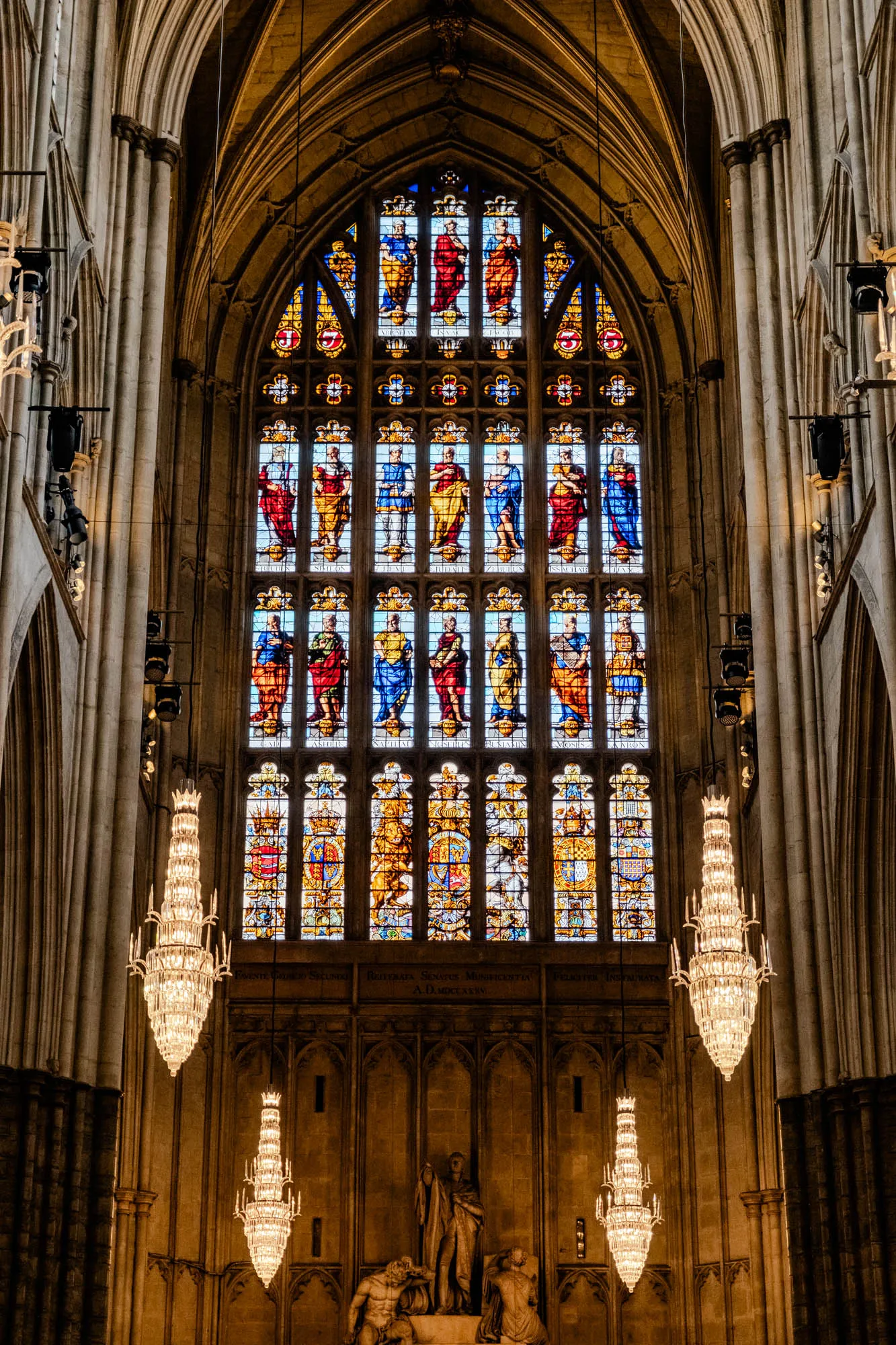 The image shows the interior of a large church with a stained glass window spanning the width of the wall. The window is made up of many small panes, each containing a different scene. The window is framed by large, arched columns that rise up to the ceiling. There are two ornate chandeliers hanging from the ceiling on either side of the window. Below the window is a large statue of a man. The walls of the church are made of stone and the lighting is dim. The stained glass window depicts various figures, possibly religious figures, in different poses and with colorful clothes. Some text is visible in the window which likely identifies the figures.  It is possible that the church is Westminster Abbey. 
