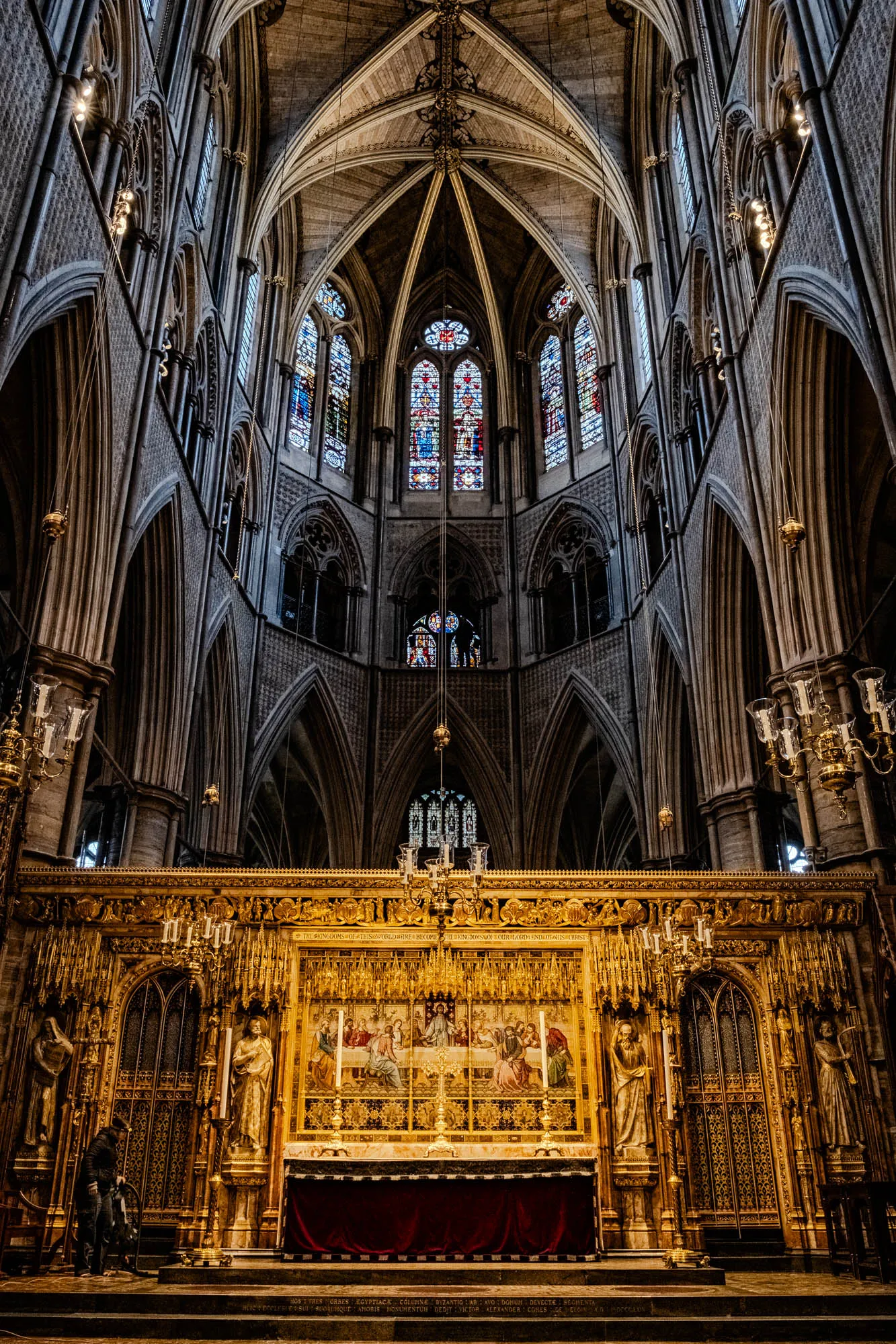 The image shows the interior of a grand cathedral. It is a high-ceilinged space with large arched windows. The windows are filled with stained glass, giving the room a colorful, mystical feel. The ceiling is also arched, with intricate details of stonework and carvings. In the foreground is a massive golden altar with an ornate design. The altar is decorated with a large painting of people gathered around a table. The altar has a red cloth placed on the surface and looks like a place for ceremonial activities. There are also several ornate, gold-painted sconces with candles that hang from the ceiling. The entire scene conveys a sense of grandeur and spirituality.