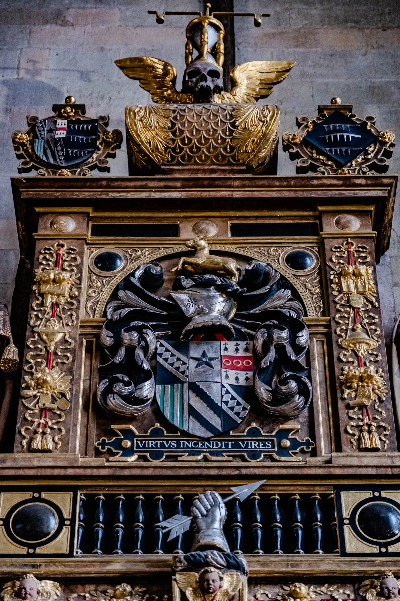 The image depicts a carved wooden monument, likely from a church. The monument is ornately decorated with gold paint and intricate details.  At the top, a skull sits within a gold and black eagle's wings, suggesting a connection to death and mortality.  Below the skull, a large wooden coat of arms is prominently displayed. The shield is divided into four sections, showcasing various symbols including a star, a checkered pattern, and a set of three rings. Above the shield, a golden deer is leaping.  On the shield's banner, the words "Virtus Incedit Vires" are visible, which translates to "Valor Kindles Strength." 

The monument's lower section features a black, wooden grate with a series of vertical spindles.  An armored hand emerges from behind the grate, holding a silver arrow.   Two cherub statues are visible below the hand, suggesting a connection to innocence and eternity.  The entire monument is richly decorated and exudes a sense of history, faith, and nobility. 
