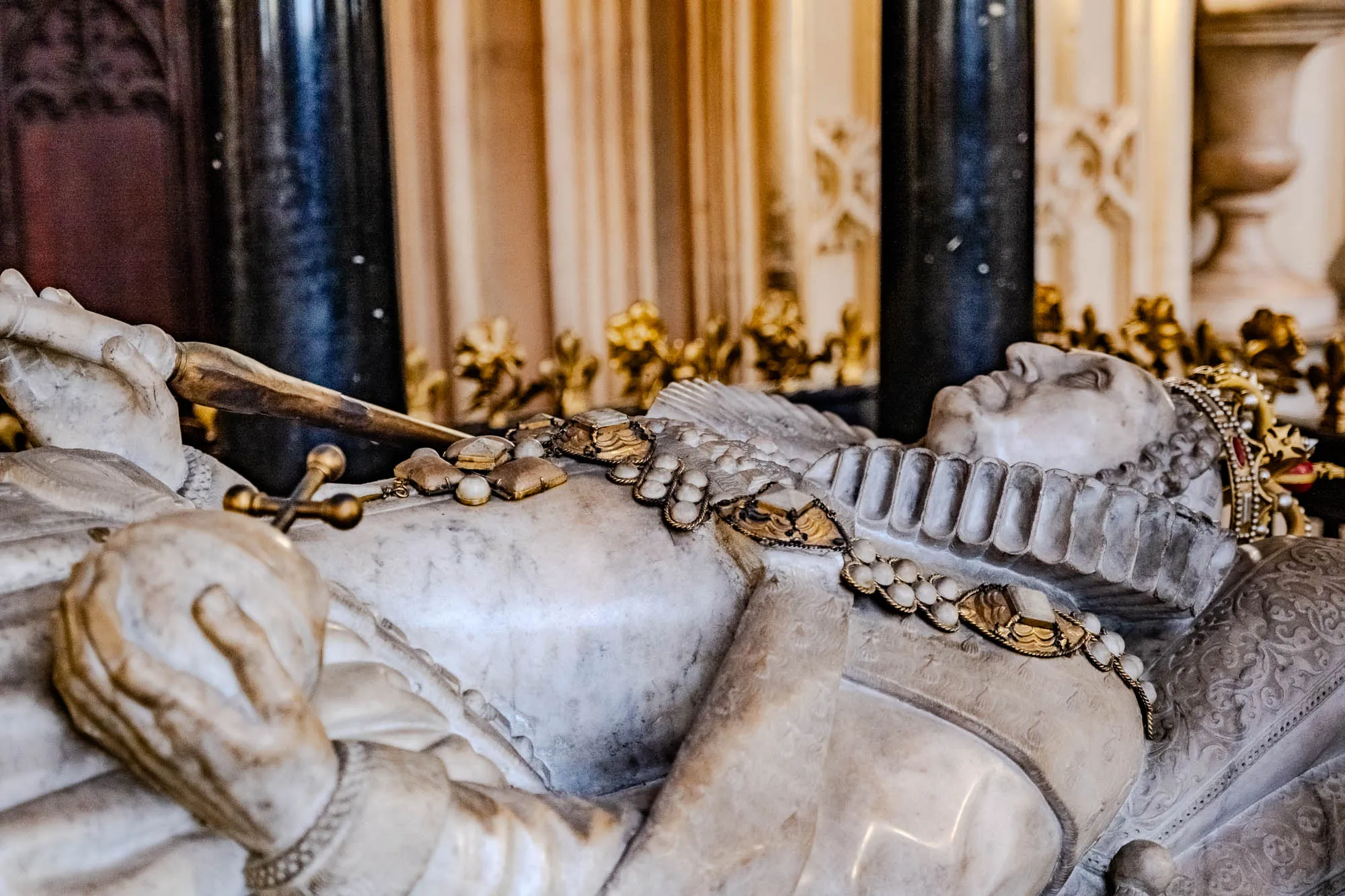 The image shows a marble effigy of a person lying on their back. The effigy is wearing a crown and is adorned with gold jewelry. The person is lying on their right side, their left hand is resting on their chest and the other hand is holding a sword. The effigy is located inside of a building, and behind the effigy, there is a tall, black pillar and a cream-colored wall with some decorative detail. The floor of the building is not visible in the image.