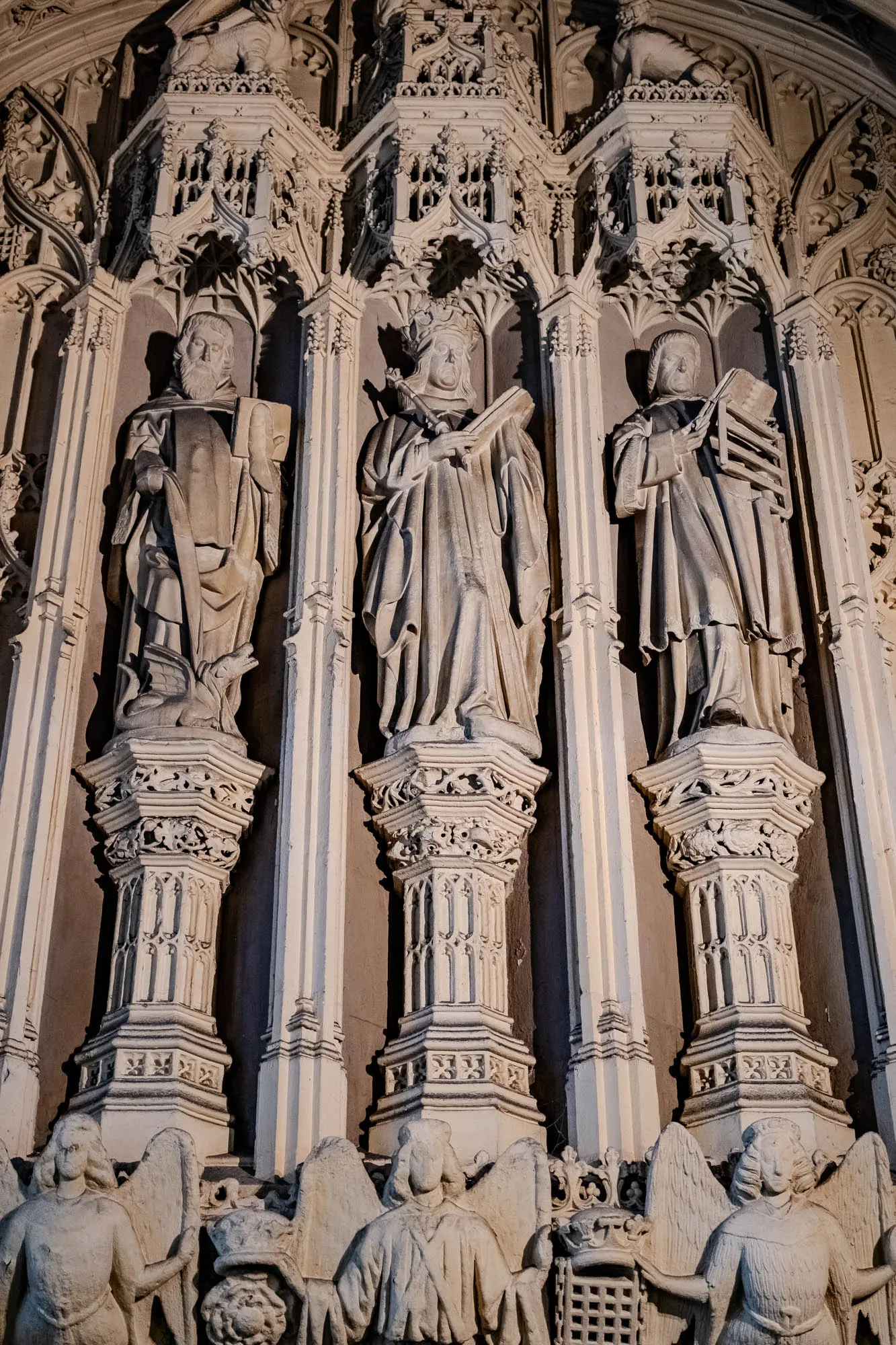 The image shows a section of a church interior with three stone statues of figures wearing robes, standing in niches.  The figures are each holding a book. All three are standing on top of decorative stone columns that feature intricate designs. There are also smaller statues of angels, carved on either side of the three main statues. The background is the stone wall of the church, which appears to be ornate with similar intricate stone carving.