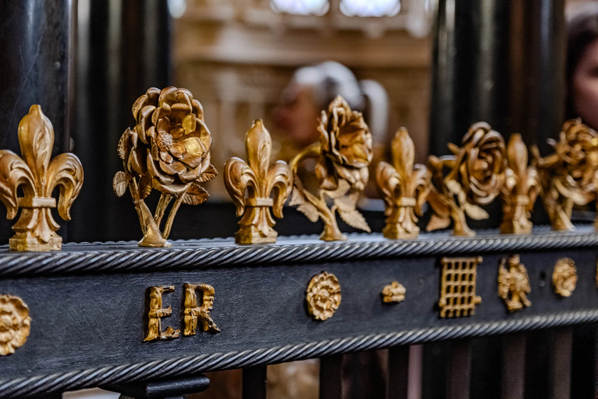 The image is of a black railing with ornate golden embellishments. There are six decorative golden roses, each with a stem and leaves. The roses alternate with golden fleur-de-lis, a stylized lily. The railing has a black, rope-like trim. On the top surface of the railing, there is a golden inscription that reads "ER," for "Elizabeth Regina," or Queen Elizabeth. There are also a variety of other designs on the surface of the railing, including a gold rose, a crown, a stylized grid-like structure, and a design that looks like a figure holding something. The background of the image is a blurred, out-of-focus scene inside Queen Elizabeth's tomb.
