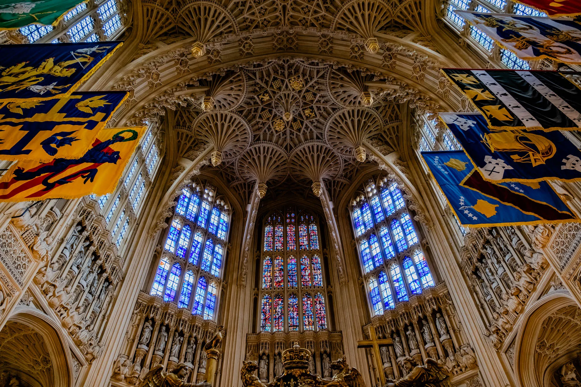 The image is a low-angle view of the interior of a large, ornate cathedral.  The ceiling is vaulted and made of stone, with many intricate carvings and details. The walls are also made of stone, and they are lined with tall, stained-glass windows. The windows are primarily blue in color, with some red and yellow hues. In the center of the image, a large stained-glass window dominates the space. It is made of a large number of smaller panes, each with a different color or design. The window is surrounded by a series of white columns. Along the walls of the cathedral, there are many heraldic banners hanging from the ceiling. The banners are made of colorful fabric with intricate designs, often featuring coats of arms. These banners are hanging from the ceiling, and they are illuminated by the light coming through the windows. The banners are tilted, as if blown by a gentle breeze. There are also many sculpted figures on the walls, creating a sense of religious reverence and history. The image is captured from a low angle, making the cathedral seem even grander and more imposing. The light filtering through the stained-glass windows creates a colorful and mystical atmosphere, adding to the image's overall sense of awe and wonder. The scene feels quiet and peaceful.