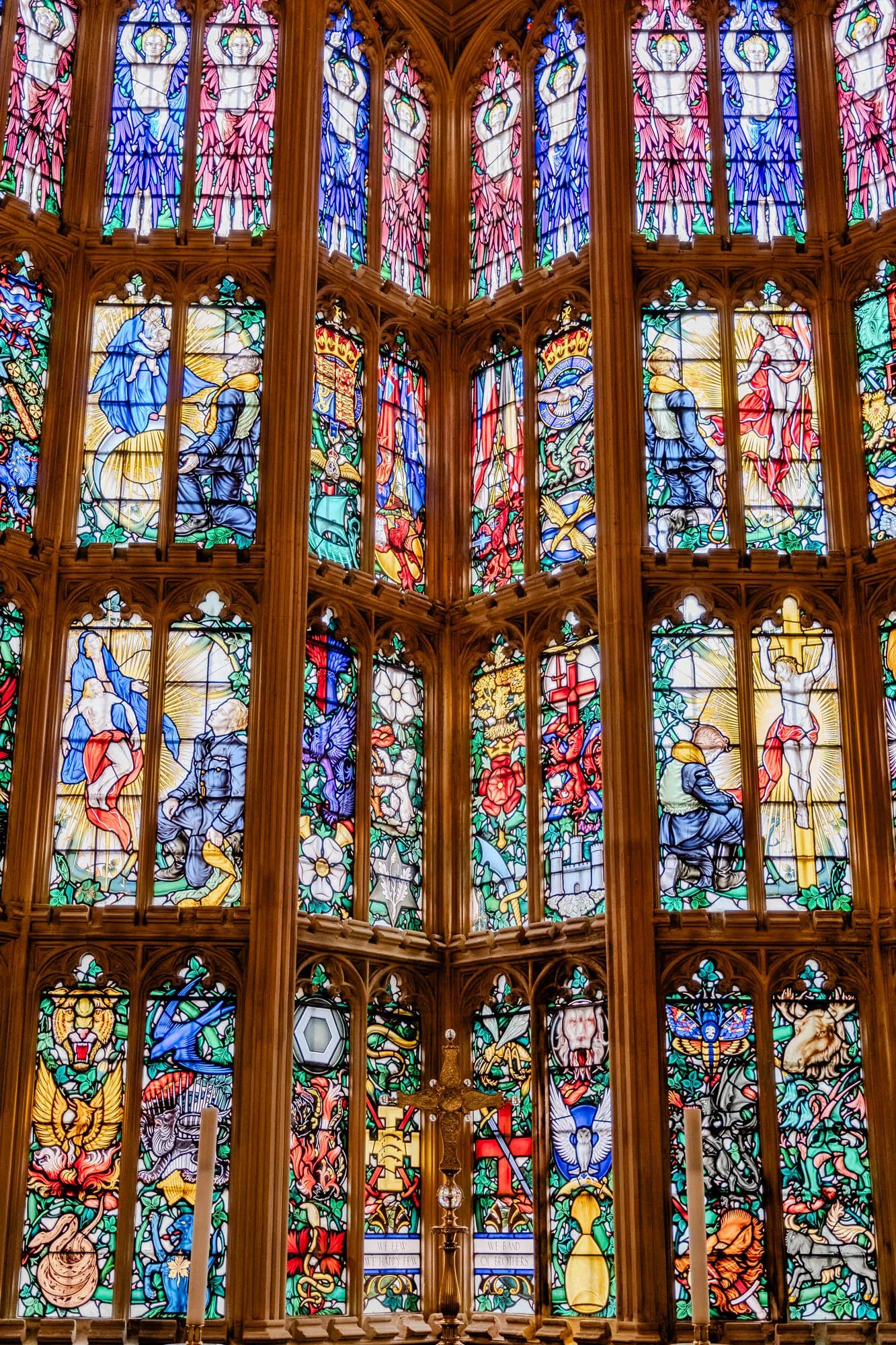 This is a stained-glass window composed of many individual panes of glass. The window is shaped like a triangle, with a narrow point at the top and a wide base at the bottom. It is divided into 15 separate sections, each containing a different image.  

The top row features angels with outstretched wings. In the middle row, some panes show figures praying or kneeling, while others depict heraldry, including a shield with a cross on it. 

The bottom row features various colorful images, including a lion, a bird, a bee, an owl, a moth, a deer, a dragon, a sword, a cross, a shield, and a snake.

The entire window is bordered by a wooden frame, and there is a golden cross in the center of the window.  The stained glass glows in the light and makes a beautiful pattern of color.