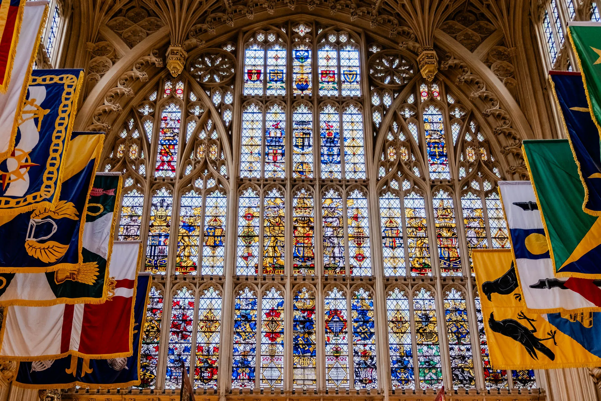 The image shows a large stained glass window in a stone archway. The window is divided into many panes, and each pane contains a different coat of arms. The window is bathed in light, and the colors are bright and vibrant. On either side of the window, there are several flags hanging, with colors of white, blue, yellow, green, and red. The flags are fringed with gold, and they are arranged in a way that suggests they are hanging in a formal setting. The room appears to be a historic building, possibly a chapel or church.  The image evokes a sense of grandeur and history.