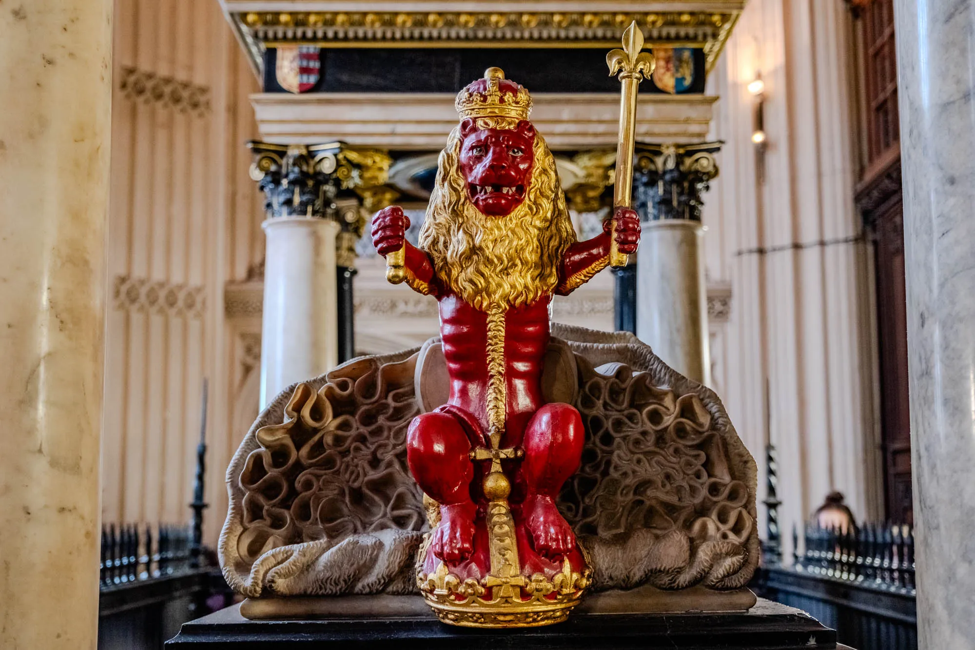 The image shows a statue of a red lion in a seated position. The lion has a gold crown on its head and its mane is also made of gold. It is holding a gold staff with a fleur-de-lis on top.  The lion is seated on a gold and white ornate base. The lion's body is painted red and the background is out of focus but appears to be the interior of an old building.  The image appears to be part of a larger monument.