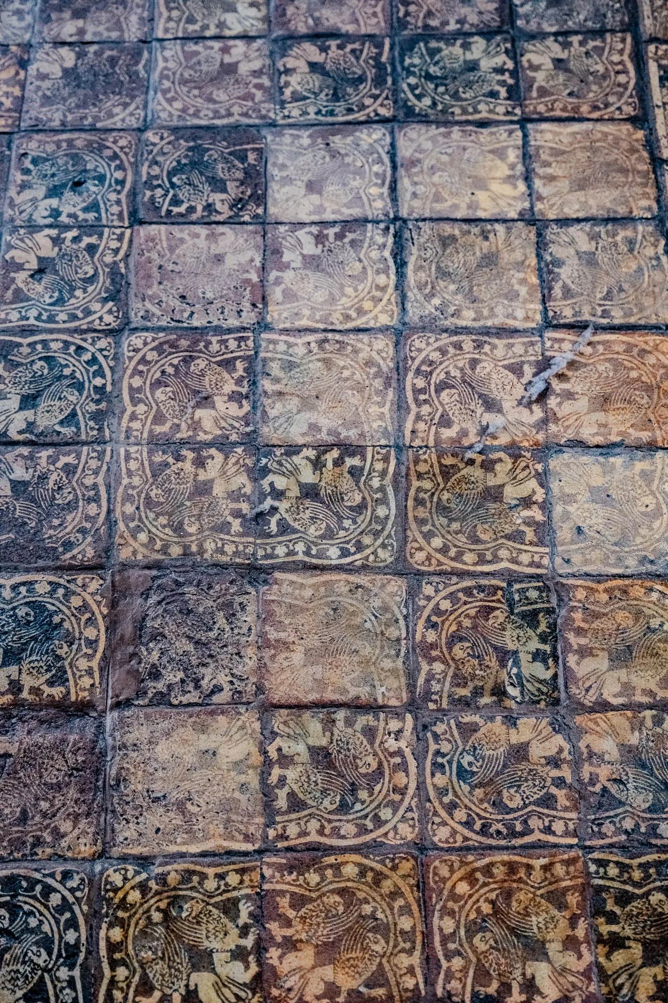 This is a close-up image of a tiled floor. The tiles are square and are arranged in a checkered pattern. The tiles are made of a dark brown material and have a faded, light brown design on them. The design is an intricate pattern of leaves and small birds. Some of the tiles are more faded than others.  The tiles are worn and some of the edges are chipped. The tiles are arranged in a grid pattern and are close together.