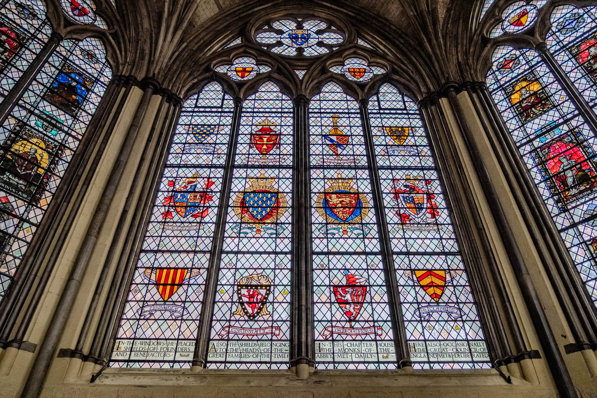 This image is a close up of a large stained glass window in a stone cathedral. The window is divided into 10 sections and each section contains a shield with a coat of arms. Each shield has a unique pattern and color of red, yellow, blue and gold. Each shield is surrounded by a blue and white lattice pattern and has text written on the bottom. Below the bottom row of sections there are three signs with text. The first sign says "IN THE WINDOWS HE PLACED SHIELDS OF FOUNDERS AND BENEFACTORS," the second sign says "ALSO THE SHIELDS BELONGING TO THE HEADS OF THE GREAT FAMILIES OF THE TIME", and the third sign says "IN THIS CHAPTER HOUSE THE MONKS OF THE ABBEY MET DAILY HERE ALSO OCCASIONALLY THE GREAT COUNCIL OF ENGLAND MET & DELIBERATED". 

The overall tone of the image is historic, beautiful, and intricate.  