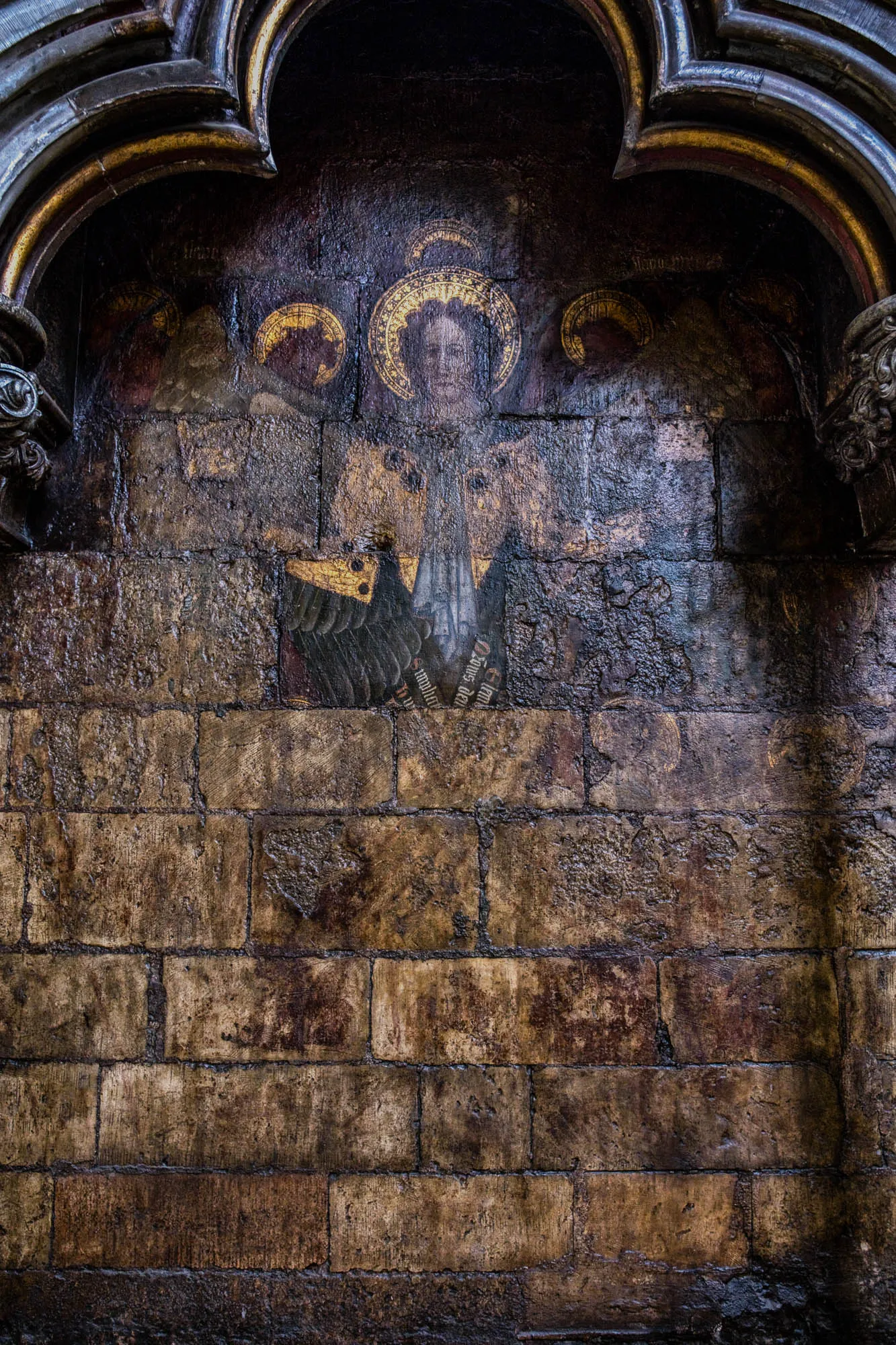 A close up of an old stone archway with a faded painting of a person with wings. The painting is in the center of the arch, and is mostly obscured by the dark stone of the arch and the weathering of the stone wall below it.  The painting appears to be of a figure with large wings spread, and a halo, possibly an angel.  There is some inscription on the wall to the right of the figure. The stone archway has a golden trim. The stone wall below the arch is made of large, rectangular stones that are brown, gray, and black.  The wall is worn and weathered, with some staining from water damage. The image may be of the interior of an old church.