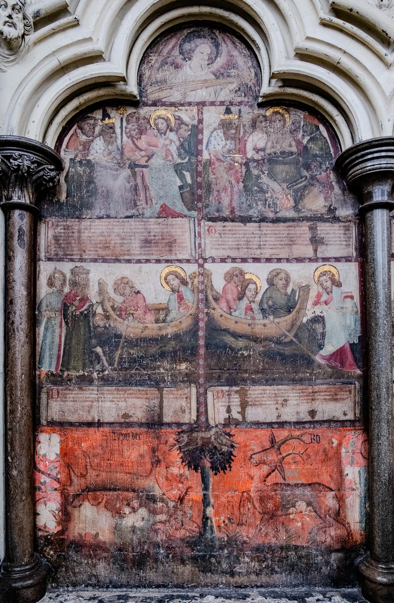 The image depicts a faded fresco painting, divided into five sections. 

The top section shows a figure with halo, lying on a bed, surrounded by four figures.  They are likely meant to represent a biblical scene of a religious figure or a saint.

The second and third sections depict a scene where seven figures are sitting inside two boats on top of a dark and choppy water. They are likely meant to represent a biblical scene of a religious figure or a saint. 

The fourth section appears to be a scene with a large, brown deer, standing in a field of green grass.  The top of the deer's antlers are broken off. To the right of the deer, is a partial figure of a person in a robe, standing to the right of the deer.

The fifth and bottom section is a landscape with a tree growing on the left side of the image.  The tree is a dark brown color, and the foliage is dark green.

All sections of the image have faded text and are in the style of medieval paintings, but they appear very old and heavily damaged.  The image appears to be on a stone wall.