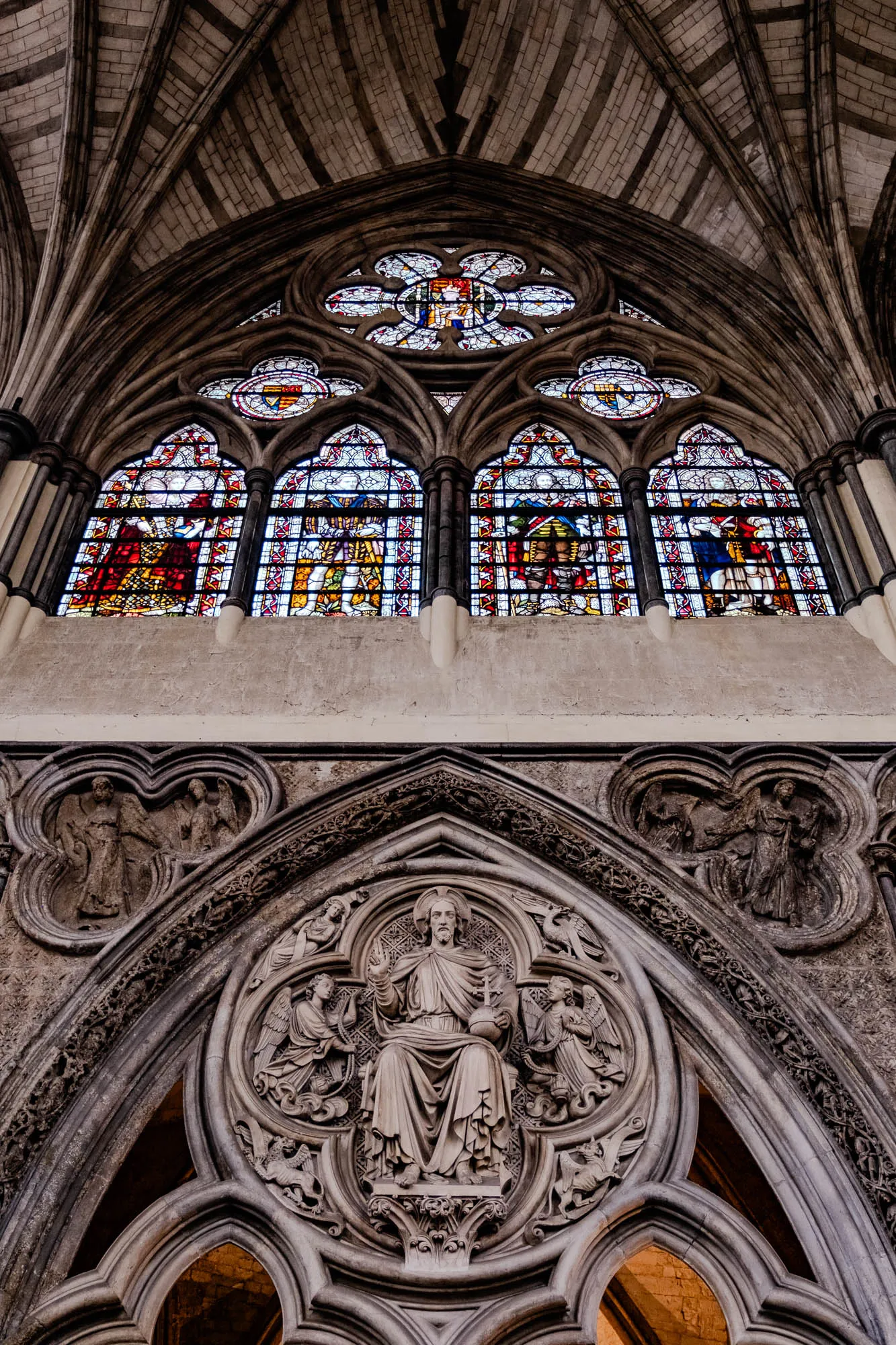 The image shows a vaulted ceiling with a large stained glass window in the back and an ornate stone carving in the foreground. The stained glass window has four panels depicting people in robes. The window is set in an arched stone frame and is framed on either side by columns. The vaulted ceiling is made of stone blocks and has a series of arched ribs running along the length of the ceiling. The stone carving is circular and depicts a man with a beard sitting on a throne. He is surrounded by angels and other figures. The carving is set in a round arch and is surrounded by intricate carvings of flowers and other patterns. The overall effect of the image is one of grandeur and beauty.